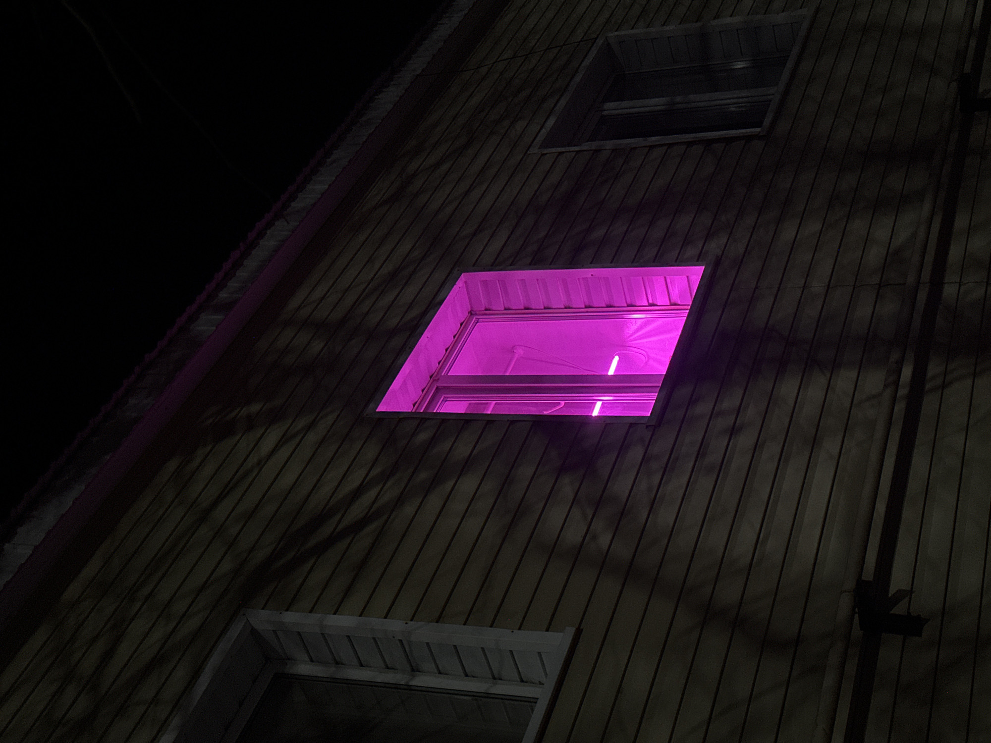 A low-angle shot of the side of a dark, three-story building at night. The exterior is comprised of vertical wood panels. The top and bottom windows are dark, while the middle window is brightly illuminated in pink. A downspout and other fixtures run down the right side of the building. Shadows cast on the building from trees. The location is presumably Aktanysh, Russia.