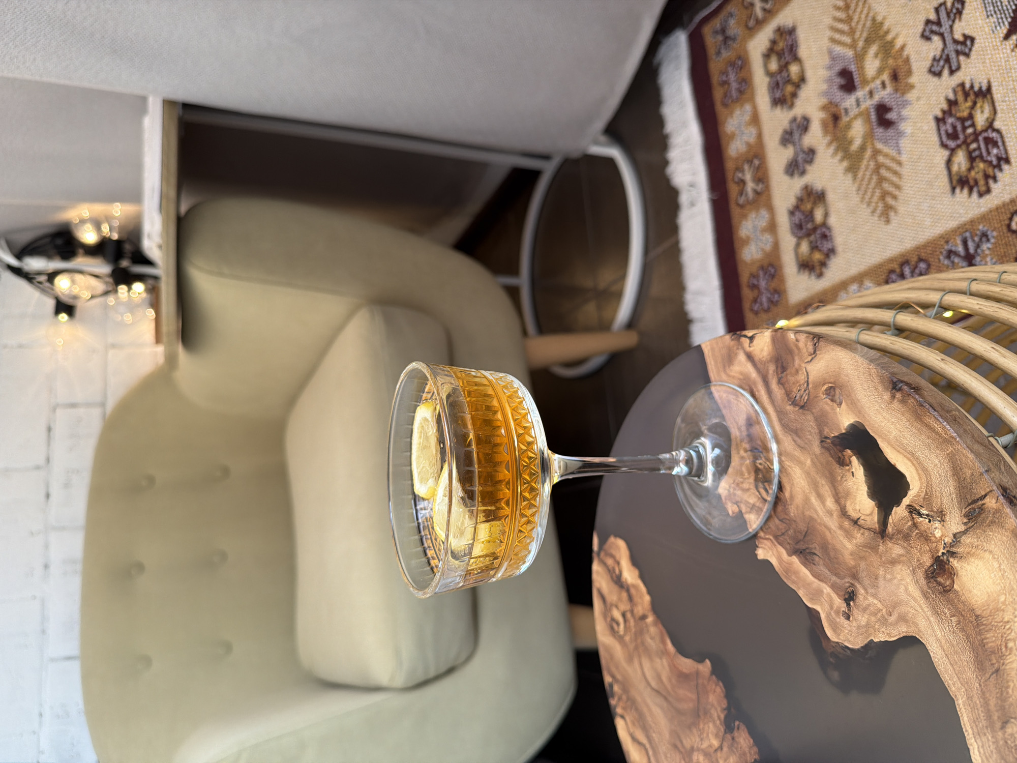 The photograph presents a close-up, top-down view of an interior scene. A light green upholstered armchair is positioned in the lower-left quadrant. Next to the armchair, a cocktail glass filled with a yellow liquid, possibly a cocktail, and lemon slices rests on a round wooden table with a dark resin fill. The table's surface features intricate wood grain patterns. In the upper-left corner, a modern chandelier hangs from the ceiling. The upper-right corner reveals a patterned rug with geometric designs in earth tones. Beneath the armchair, a portion of a metal table base is visible. The room's lighting is soft and natural.
