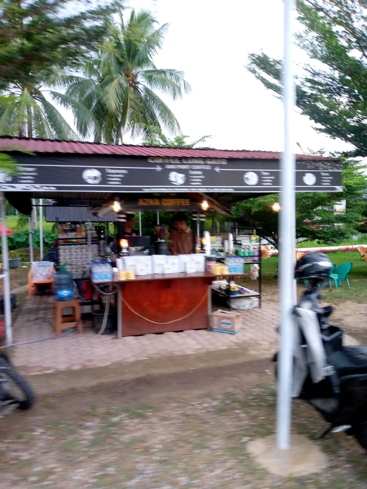A small outdoor coffee stand is set up under a red awning. The stand has a dark brown counter displaying bags of coffee and a blender. Behind the counter, two men are visible, one standing and one seated. Various bottles of drinks and snacks are displayed on shelves to the left. A wooden stool with a water cooler on top is positioned in front of the shelves. A cardboard box rests on the ground in front of the counter. Several chairs, some green and one orange, are scattered around the area. A motorcycle with a helmet on the seat is parked to the right. Palm trees and other greenery form the background. The scene appears to be in Glokaron, Indonesia.
