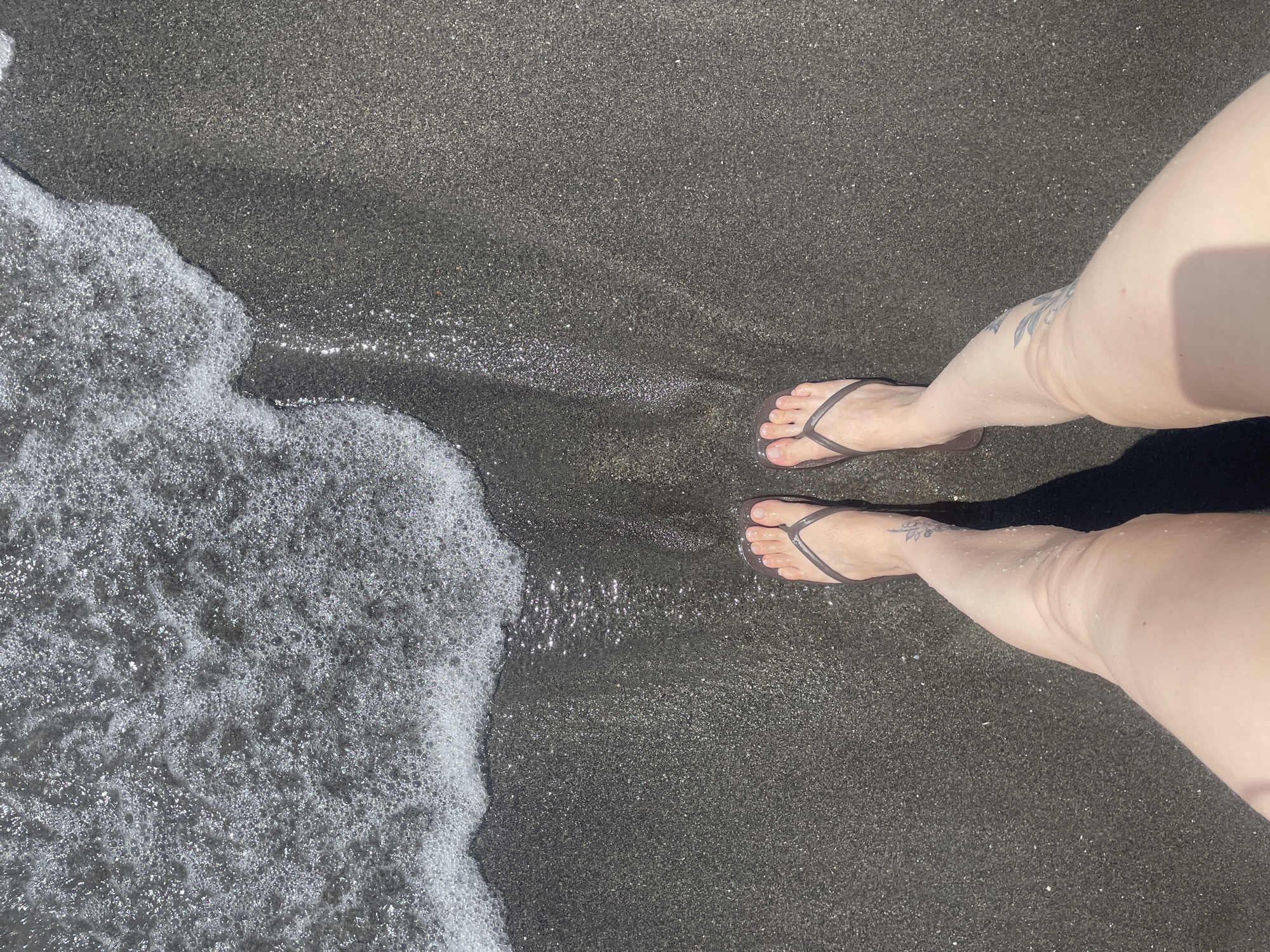 This is a high-angle, medium shot of a person's legs and feet standing on a dark sand beach at the edge of the water. The person is wearing dark grey flip-flops and has a tattoo of a floral design on their left shin. The water is shallow and foamy, with small bubbles and sparkles from the sunlight. The sand is dark and appears to be wet near the water's edge, with a fine-grained texture. The lighting suggests it is a sunny day. The image does not provide specific clues to identify the exact location within Almuñécar, Spain, beyond it being a beach environment.