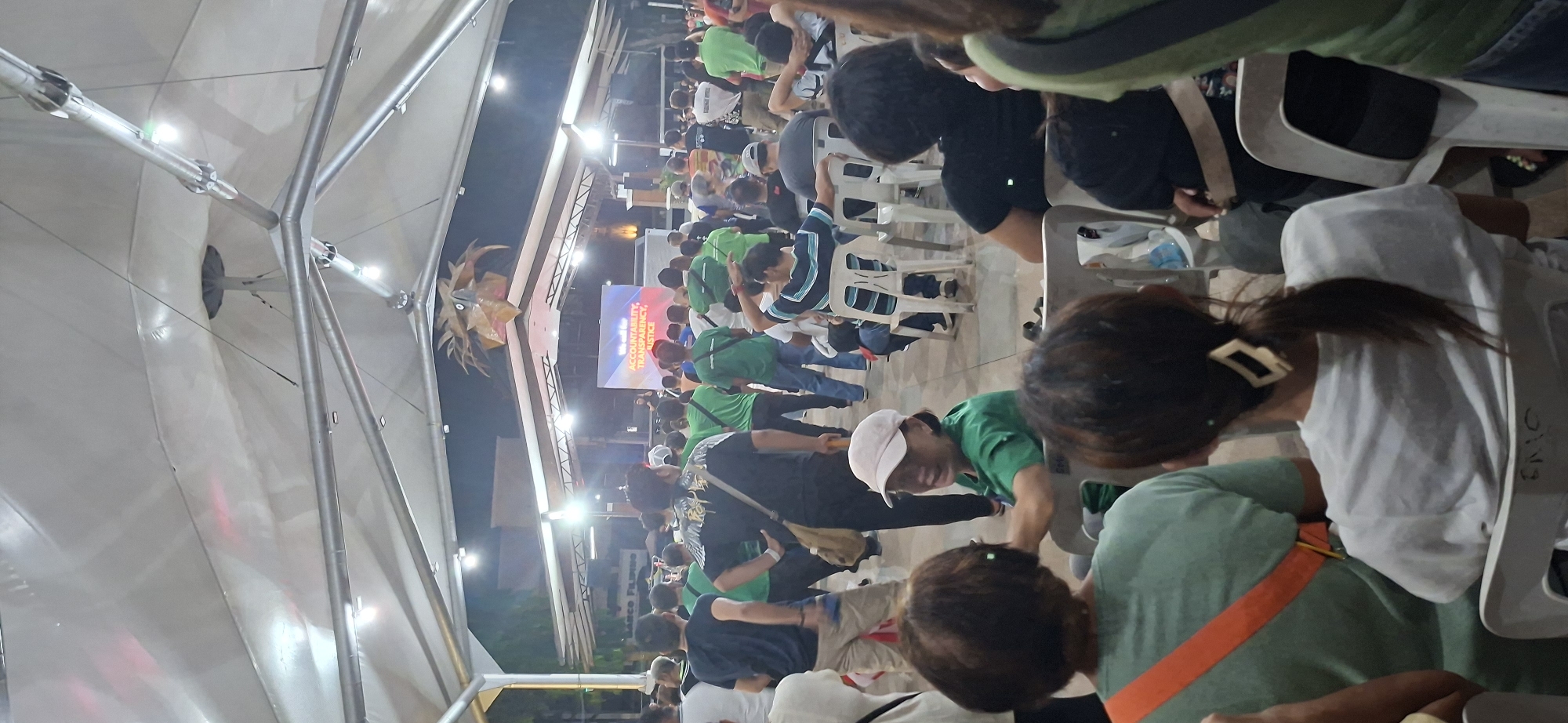This is a medium-shot, vertical, indoor photograph taken at night. The scene appears to be an outdoor event, likely a rally or a public gathering, under a large, white tent structure. In the background, a stage is visible with a large screen displaying the words "WE WILL CALL FOR ACCOUNTABILITY, TRANSPARENCY, JUSTICE" in English. A mascot, resembling a bird's head with golden feathers, is mounted above the stage.
The foreground and midground are populated with many individuals, mostly facing the stage. Many people are standing, while others are seated on white plastic chairs. The majority of the attendees are wearing green shirts. One woman in the mid-foreground, wearing a green shirt and a light-colored baseball cap, is looking back and smiling at someone out of frame. Another woman in the foreground, wearing a green shirt with an orange strap, is facing away from the camera. A person in the mid-ground is wearing a black shirt with a tan strap over their shoulder, and another person in the mid-ground is wearing a blue and white striped shirt.
The overall atmosphere suggests a political or civic event. Based on the screen text and the presence of a crowd, this event is likely taking place in a public space within Davao City, Philippines, where such rallies are common. The tent structure and lighting suggest a temporary setup for an evening event.