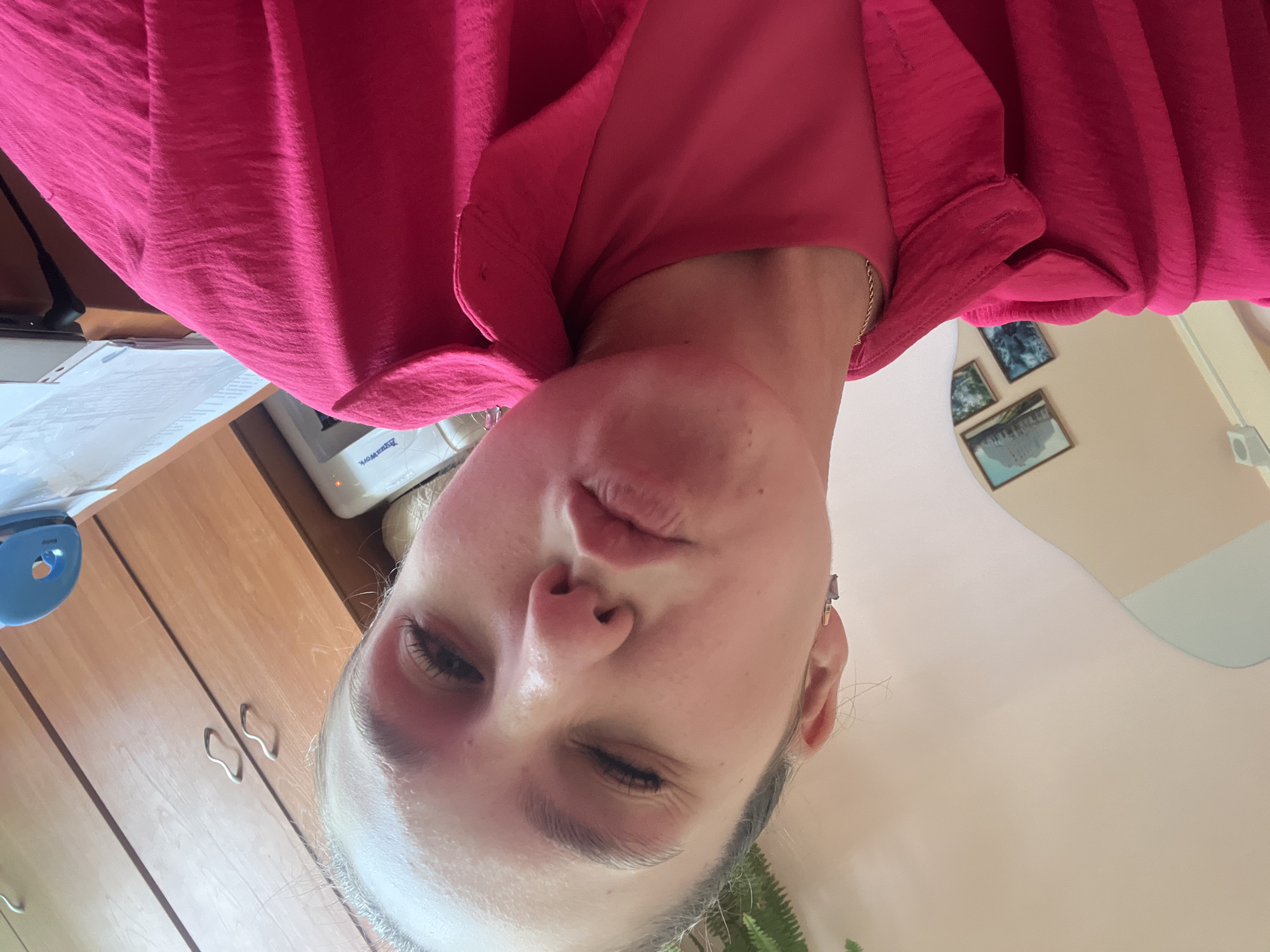 This is a selfie taken by a young person, likely female, in an indoor setting. The image is oriented sideways. The subject is wearing a bright pink, textured shirt with a collar. Their face is the primary focus, with their eyes open and one slightly squinted, and their lips pursed. Their expression appears playful or questioning.
In the background, a wooden cabinet with two silver handles is visible. On top of the cabinet, a white device with some text, possibly a label or brand name, is partially obscured. To the left of the subject's head, a blue object with a circular opening is also on the cabinet. Further in the background, there are framed pictures on the wall, depicting landscapes. The overall lighting suggests daytime. The environment seems to be a home or office space. No specific weather conditions can be inferred. There is no discernible text other than what might be on the white device in the background. The subject's jewelry, a small earring, is also visible.