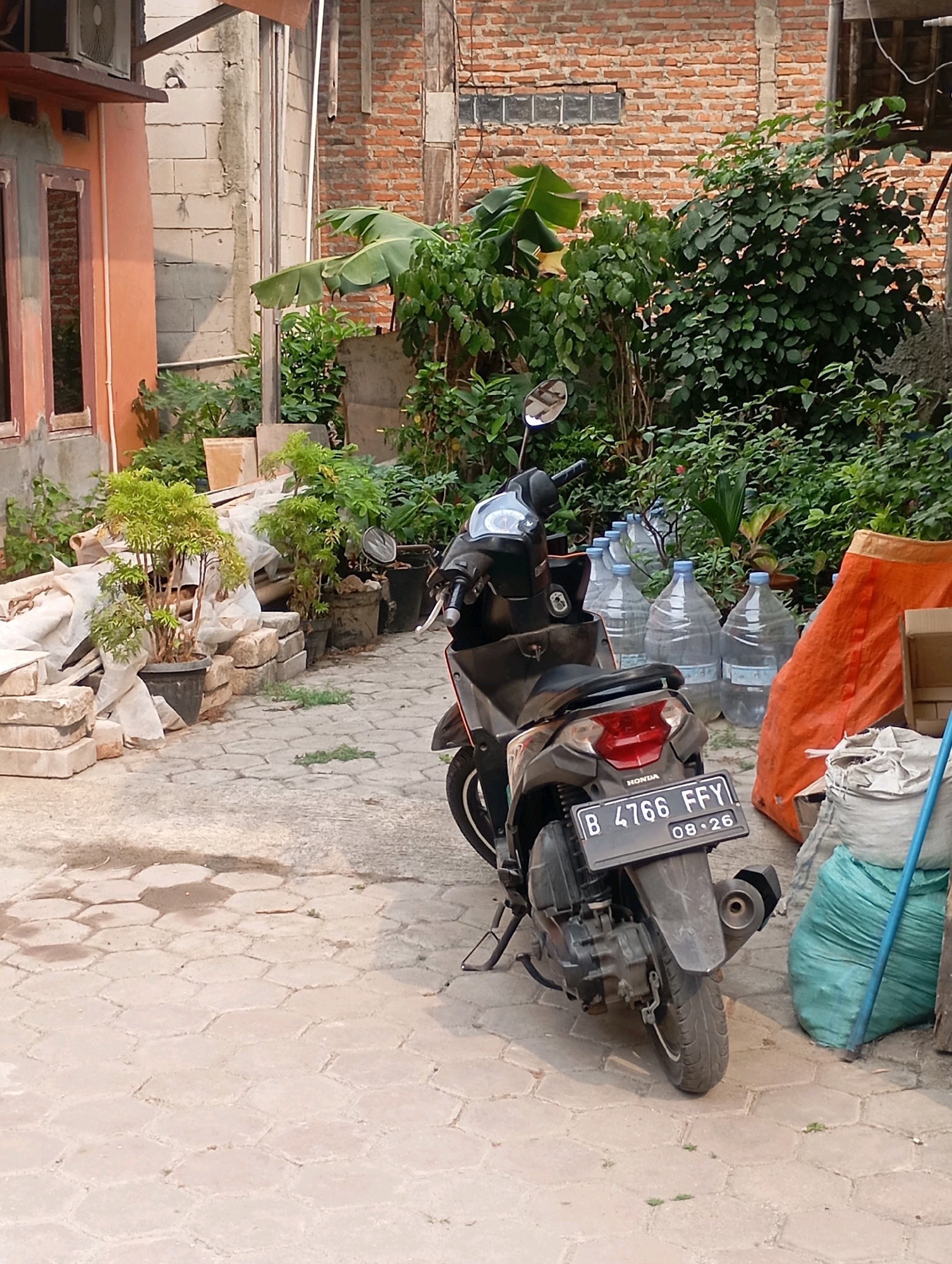 In Babelan, Indonesia, a scooter with license plate B 4766 FFY and sticker 08-26 is parked on a paved area in front of a building. The scooter is black with some silver accents. Behind the scooter, several large plastic water bottles are lined up. To the right of the water bottles, an orange tarp is visible, as well as a green bag and a blue pole. The background consists of lush green foliage and a brick building. The paving stones are hexagonal.