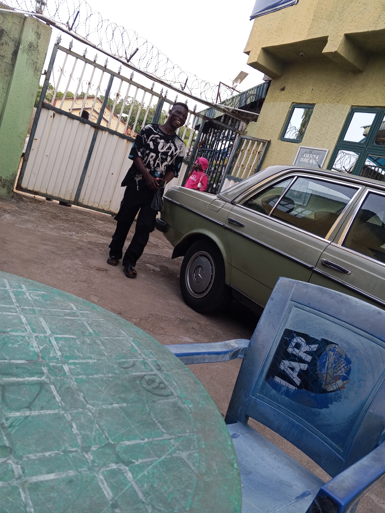 The image captures an outdoor scene, likely during daytime, in Zango-ndaji, Nigeria. In the foreground, a round, weathered green plastic table partially obscures a blue plastic chair with a torn sticker displaying "IAR".
The central subject is a young man, standing on a dirt ground and smiling widely, his mouth open in laughter or a joyful expression. He wears a black short-sleeved t-shirt with a white and blue pattern, including visible text fragments like "RAG", and dark cargo-style pants with dark sandals. He holds a small, dark bag or item in his left hand.
To his right, a light green (possibly khaki) older model sedan, resembling a Mercedes-Benz W123, is parked. Its chrome trim and wheel hubcaps are visible. Behind the car, a small child in a pink top is partially visible.
The background features a tall metal gate topped with barbed wire, providing a boundary to the property. Behind the gate, another building with a light-colored facade and dark-framed windows is visible. A small sign on this building reads "ASK FOR A DUBIC M...", likely referring to a product or service. Further in the distance through the gate, another structure with text "IC UN..." can be seen. The general ambiance suggests a casual, everyday street or compound setting under bright, clear weather conditions.
