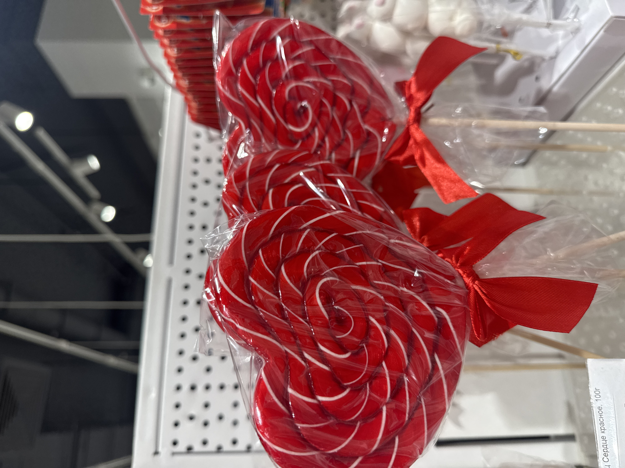 This is a close-up, eye-level shot of three red heart-shaped lollipops with white swirls, each individually wrapped in clear plastic and tied with a red ribbon. The lollipops are arranged in a row, with the leftmost one most in focus. Behind them, other candies and store shelving are visible. The overall scene suggests a retail environment, likely a confectionery or gift shop. The image is undated and unlocated, therefore I cannot provide any details about its specific location in Ufa, Russia.