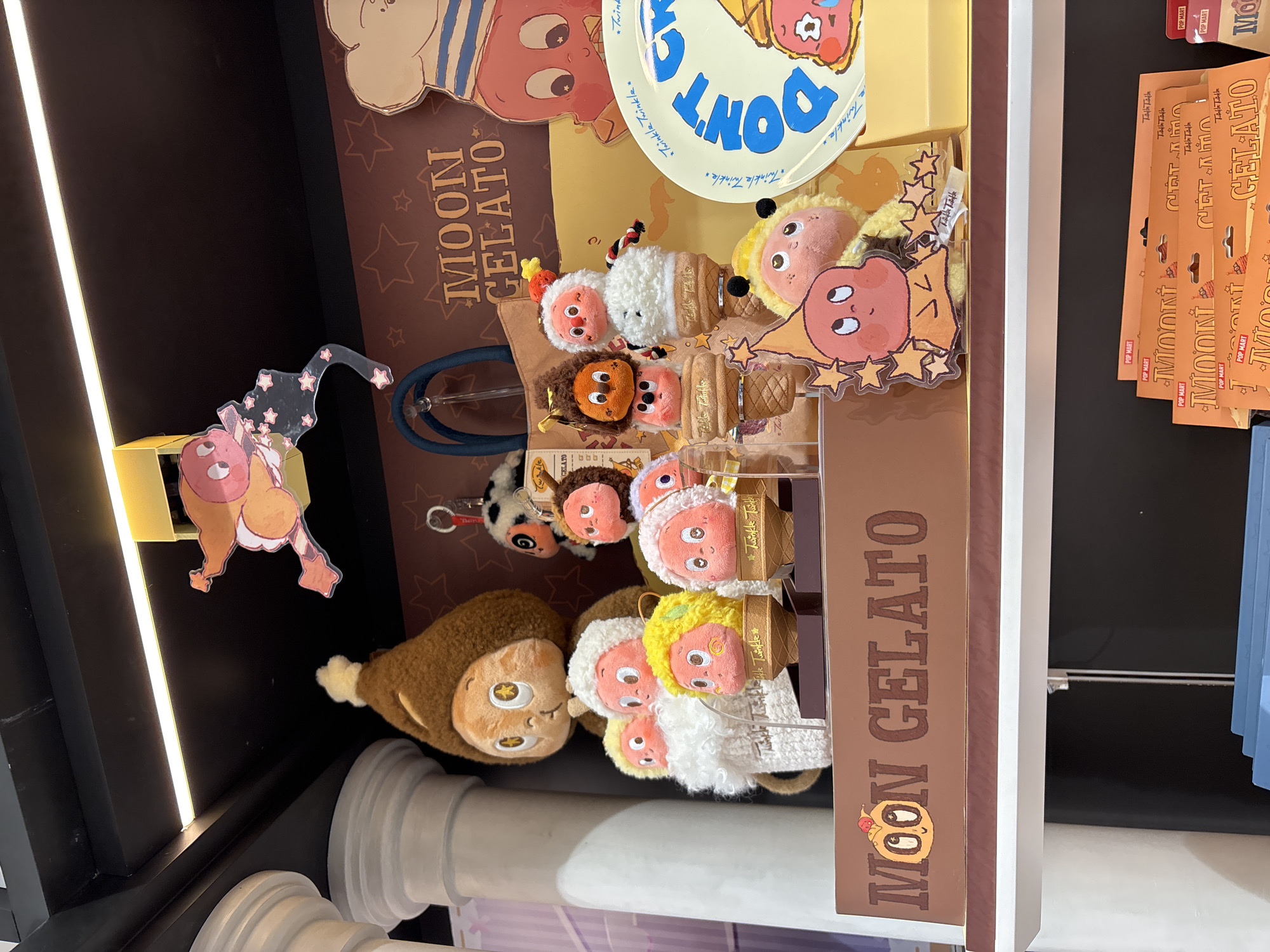 This is a photograph of a display of "Moon Gelato" branded merchandise. It's most likely taken inside a retail store, given the structured display and the presence of product packaging.
The main objects are plush toys resembling ice cream cones, each with a different color and a cartoon face. There's also a larger plush toy that appears to be a monkey or similar creature. Additionally, there are boxes and packaging visible, also branded with "Moon Gelato."
In the background, there is a display board with stars and a cartoon character. A blue hose-like object is also visible.
The photograph seems to be taken in West Jakarta, Indonesia, judging by the location information available in the prompt.