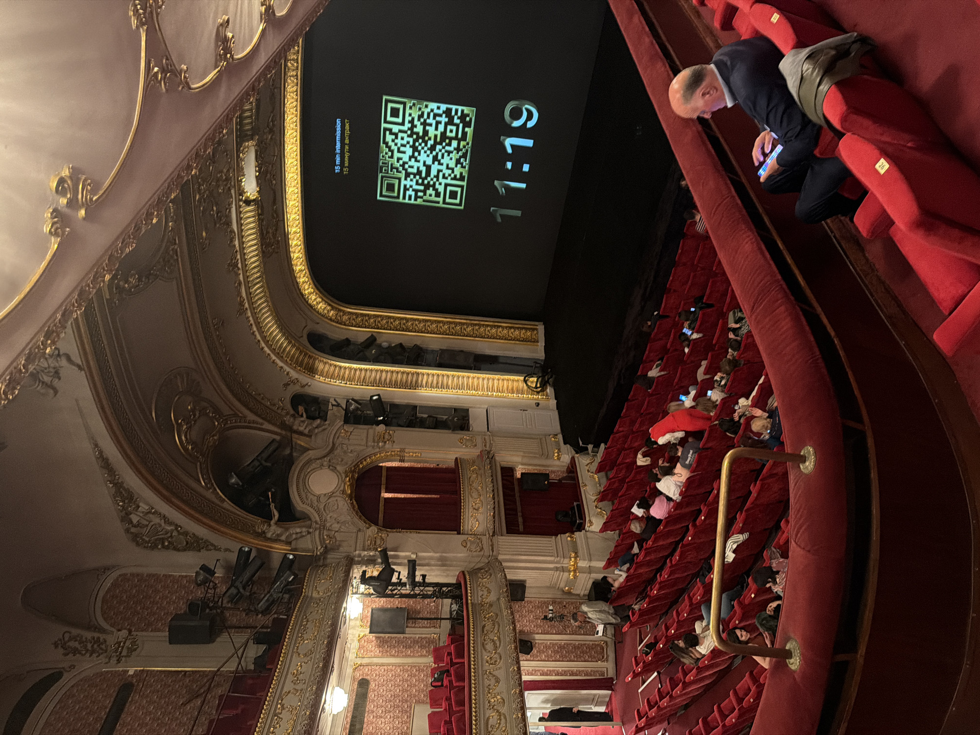 The media file captures the opulent interior of a grand theater or opera house in Sofia, Bulgaria, seen from an elevated perspective. The ornate architecture features elaborate gilded decorations on walls and ceilings, complemented by plush, red velvet seating and railings throughout the auditorium and multiple balcony tiers.
The image depicts a moment during an intermission. A large black screen at the front of the stage prominently displays a countdown timer showing "11:19," indicating 11 minutes and 19 seconds remaining in the break. Above the timer, text reads "15 min intermission" and "15 минути антракт" (Bulgarian for 15 minutes intermission), alongside a large QR code.
Numerous audience members are seated in the red chairs, many seen from behind or from a distance. Several individuals are engaged with their illuminated smartphones, a common activity during breaks. In the immediate foreground, a man in a dark suit jacket and light shirt is absorbed in his phone, with a bag placed on the adjacent seat. The ambient lighting is dim, characteristic of an auditorium during a performance break, with the screen serving as a bright focal point. Stage lights and spotlights are visible mounted on the theater's architectural elements. The scene conveys a relaxed atmosphere of waiting before the next act of a performance.