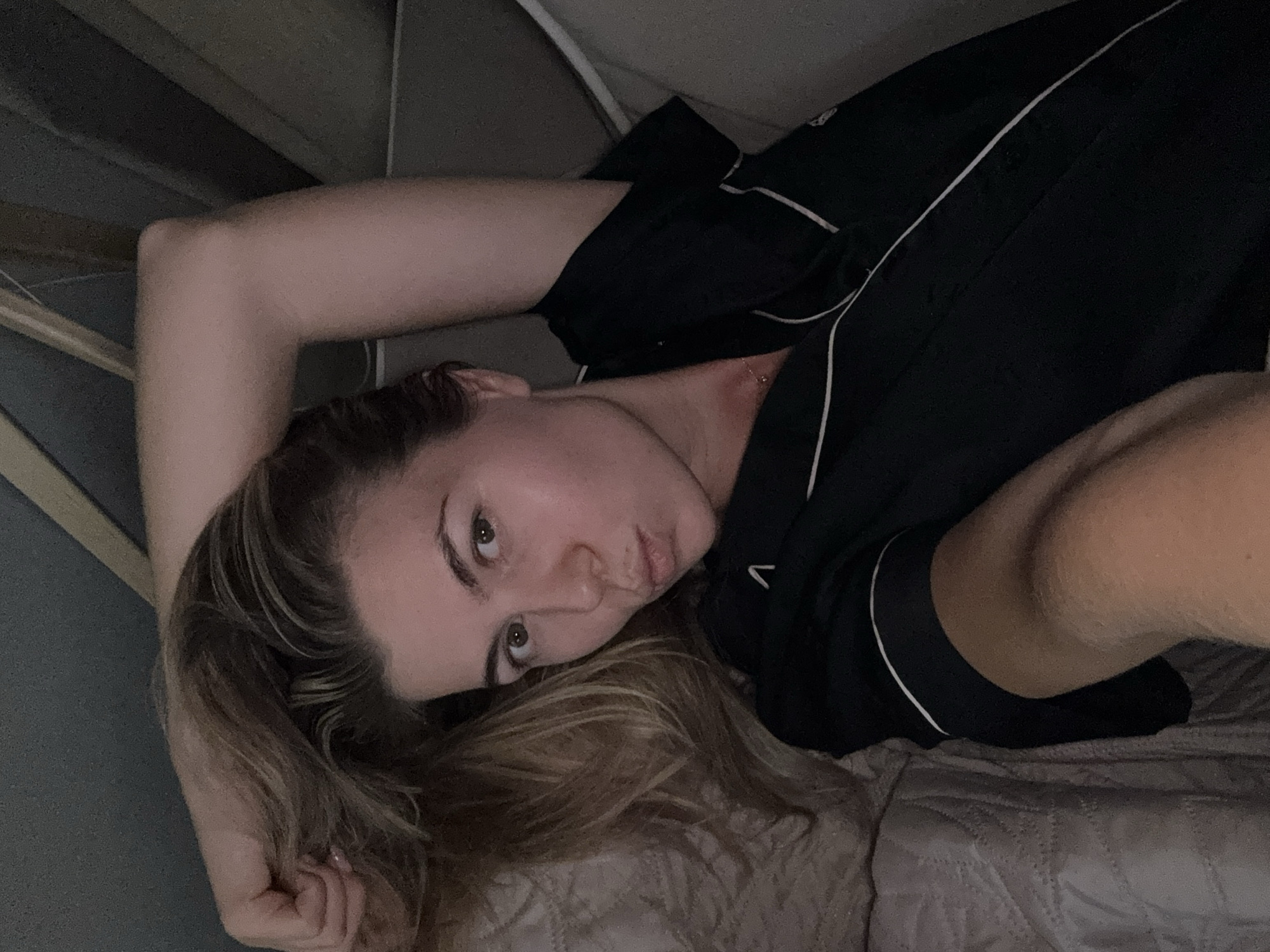 The media file is a selfie of a young woman lying on her side in bed. She is wearing black pajamas with white piping. The woman has long blonde hair and is looking at the camera with a playful expression. Her left arm is bent behind her head, and her right hand is resting near her face. The setting appears to be a bedroom, with a textured blanket or comforter visible beneath her. The lighting is dim, suggesting it is nighttime or early morning. The overall mood is relaxed and intimate. There is no visible text in the image.