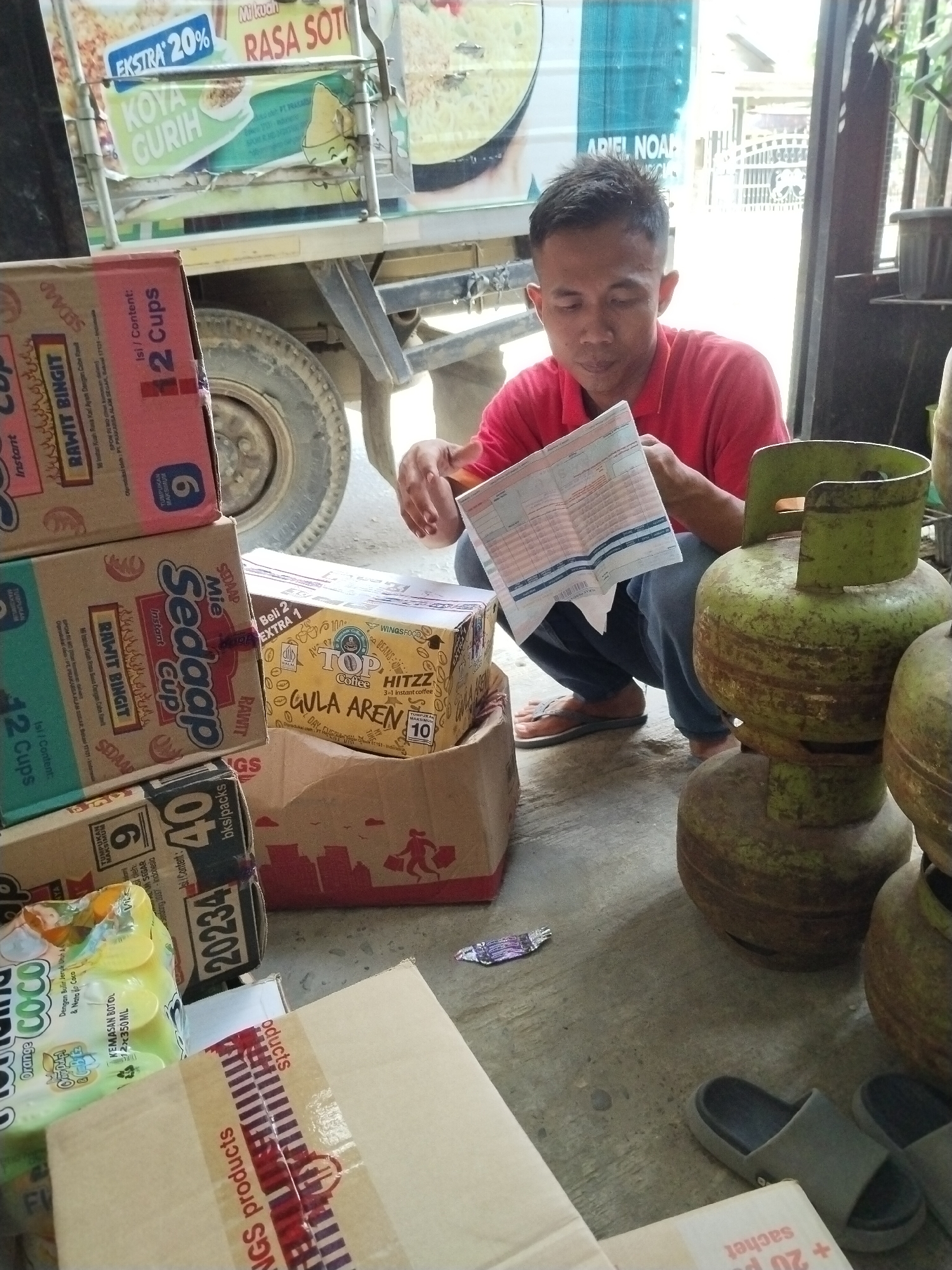 The image depicts a man squatting on a concrete floor, holding a document. He is wearing a red shirt and dark pants. In the background, a truck is visible with advertisements on its side, including text that appears to be in Indonesian. Several cardboard boxes are stacked around the man, and two green propane tanks are also present. The environment appears to be outdoors, possibly a street or an open area, with some greenery visible in the background. The man is looking down at the document he is holding. The scene suggests a commercial or logistical setting, possibly related to delivery or inventory.