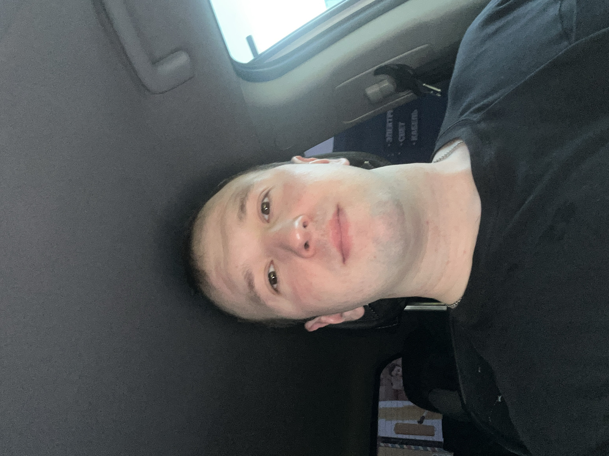 A selfie photograph shows a person in the front seat of a car. The person has light skin, short hair, and is wearing a black shirt with a silver chain visible around their neck. The background includes part of the car's interior: a gray ceiling, a window, and a black sun visor. Some text in Russian is visible on an object near the visor. It is possible this photo was taken in Chaikovsky, Russia.