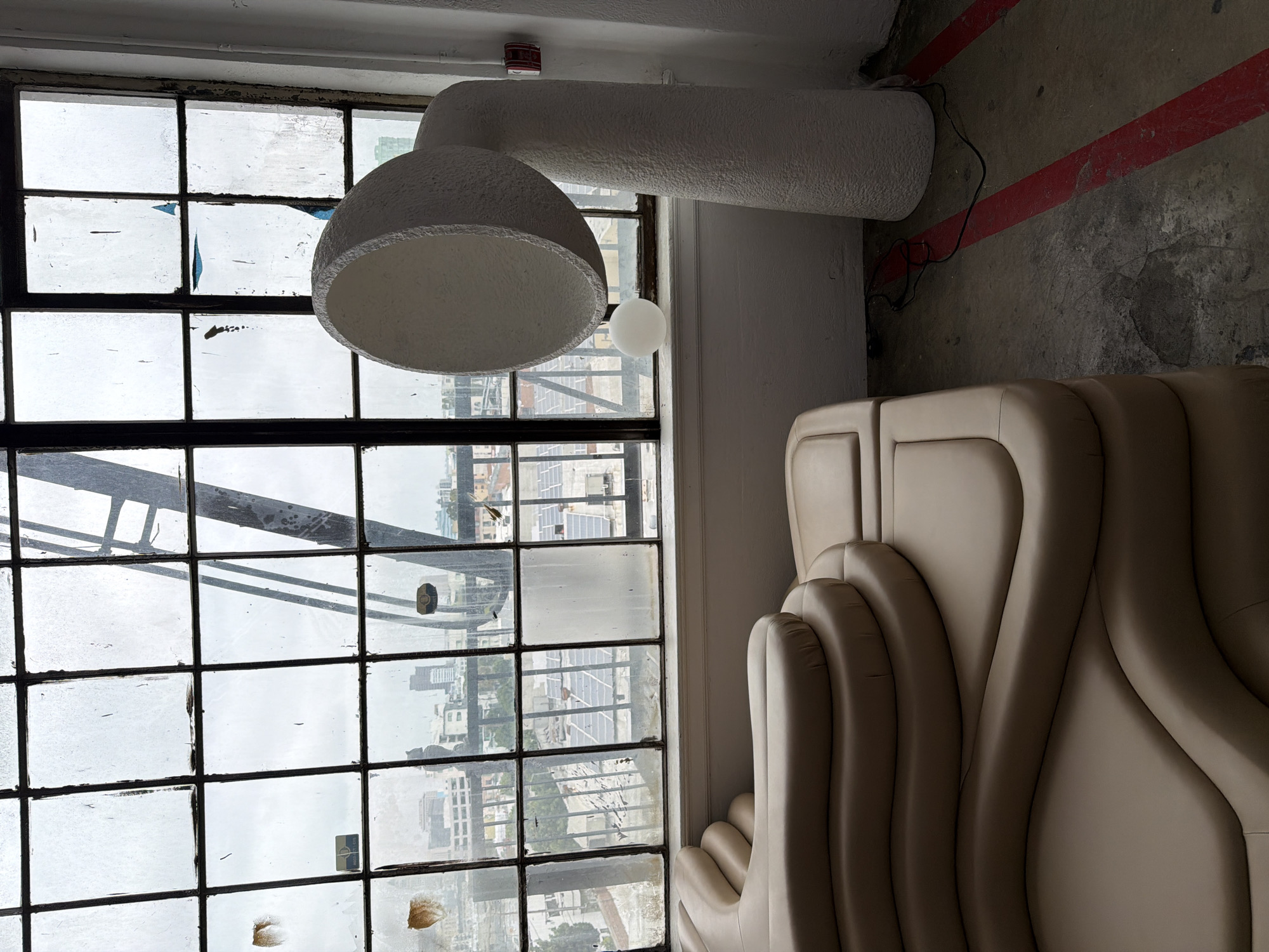 A large, irregularly shaped, light-colored, textured lamp is positioned in the right foreground of the image, adjacent to a large, multi-paned window. The window displays a city view under an overcast sky, with buildings, solar panels, and industrial structures visible. A small, white, spherical object rests on the windowsill beneath the lamp. In the left foreground, a tan, modular sofa with undulating, layered cushions occupies a significant portion of the frame. The floor is concrete with red painted lines. The environment appears to be an interior space, possibly a studio or loft, with a rugged, industrial aesthetic, evidenced by the window condition and exposed architectural elements.