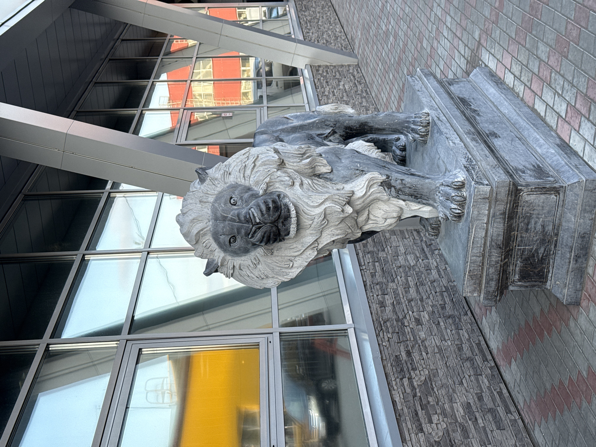 A stone lion statue stands outside a modern building in Petropavlovsk-Kamchatsky, Russia. The lion is depicted in a reclining pose on a pedestal. The building's facade features large windows with metal frames, reflecting a neighboring building. The ground is paved with both brick and stone tiles.