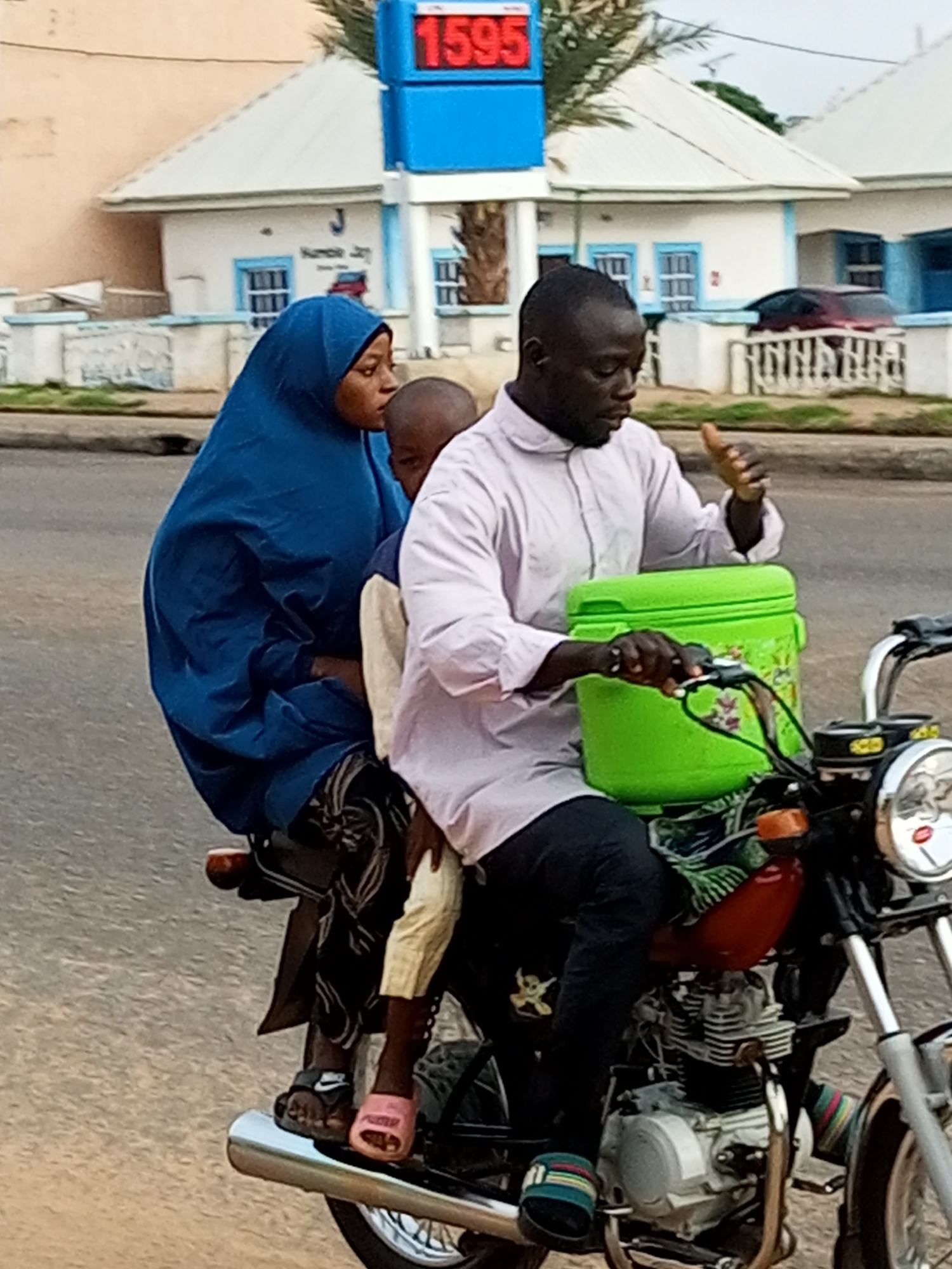 In Garko, Nigeria, three individuals are riding a motorcycle on a paved road. A man is driving, wearing a light purple collared shirt and dark pants. He is looking forward and gesturing with his right hand. To his left and slightly behind him, a woman wearing a blue hijab and patterned pants is seated, holding a child in front of her. The child is wearing a light-colored shirt and pants. A large green cooler is strapped to the back of the motorcycle, behind the driver. In the background, there are buildings with white walls and white roofs, some with blue accents around windows and doors. A blue sign with red numbers ("1595") is visible above one of the buildings, likely indicating a gas station. The sky appears overcast.