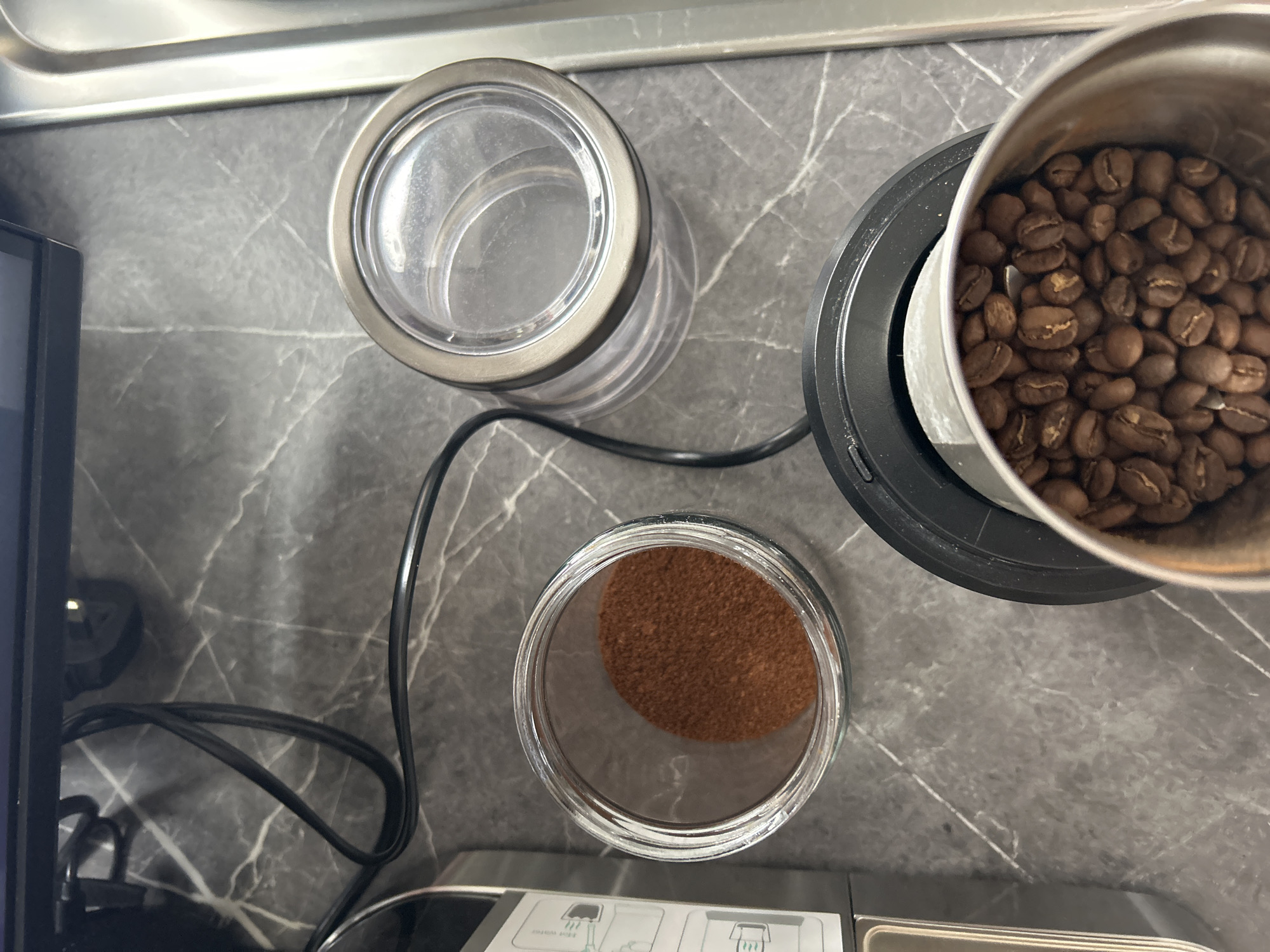 The image is an overhead shot of a kitchen countertop scene, likely indoors, with a focus on coffee-related objects. At the top right, a stainless steel coffee grinder is filled with whole coffee beans. A jar is next to the coffee grinder which appears to be empty. To the left of the grinder, a jar contains coffee grounds. In the bottom left of the shot, part of a kitchen appliance is visible, with a black electrical cord stretching out from it, near the edge of the grinder. The countertop has a grey marbled pattern. The location cannot be determined from the image.