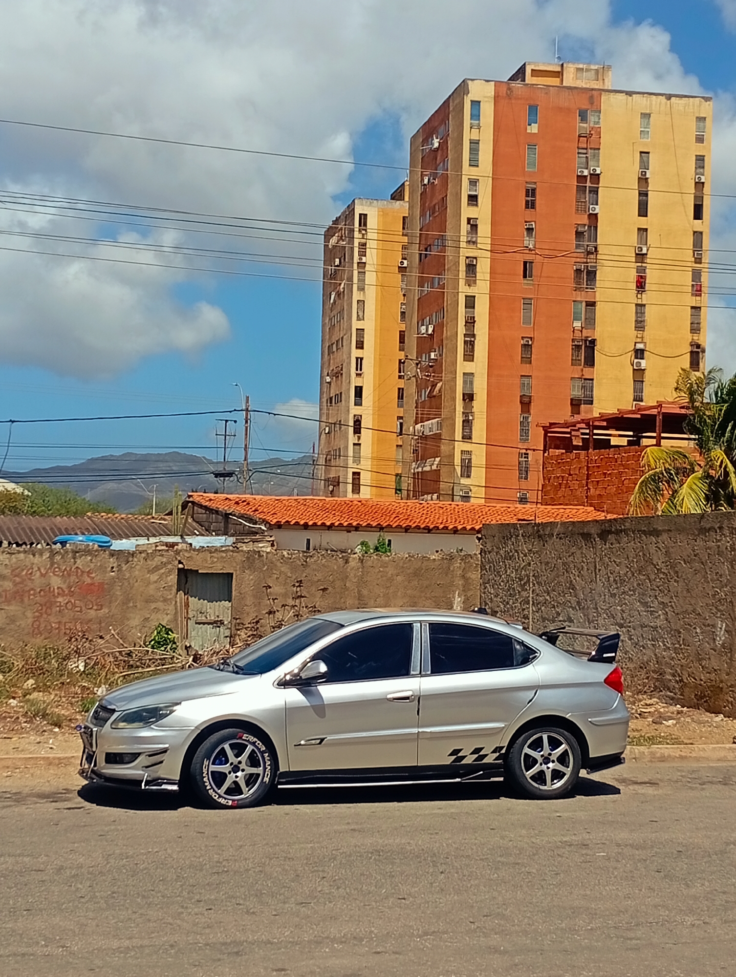 The image depicts a silver sedan parked on an asphalt road, facing left. The car has custom rims with blue accents, red lettering on the tires that reads "PERFORMANCE," and a prominent rear spoiler. Decorative checkered decals are applied to the lower side panels. In the background, two tall, multi-story buildings dominate the scene, one painted in alternating sections of orange and yellow, and the other appearing to be the same design. In front of the buildings, a low, rough-textured wall runs horizontally across the frame, with a weathered wooden door visible in its center. Beyond the wall, tiled rooftops are visible, and further in the distance, a mountain range is silhouetted against a blue sky with scattered clouds. Power lines crisscross the upper portion of the image, and a street light stands near the center of the frame. The overall environment suggests an urban or semi-urban setting. There are no people visible in the image.