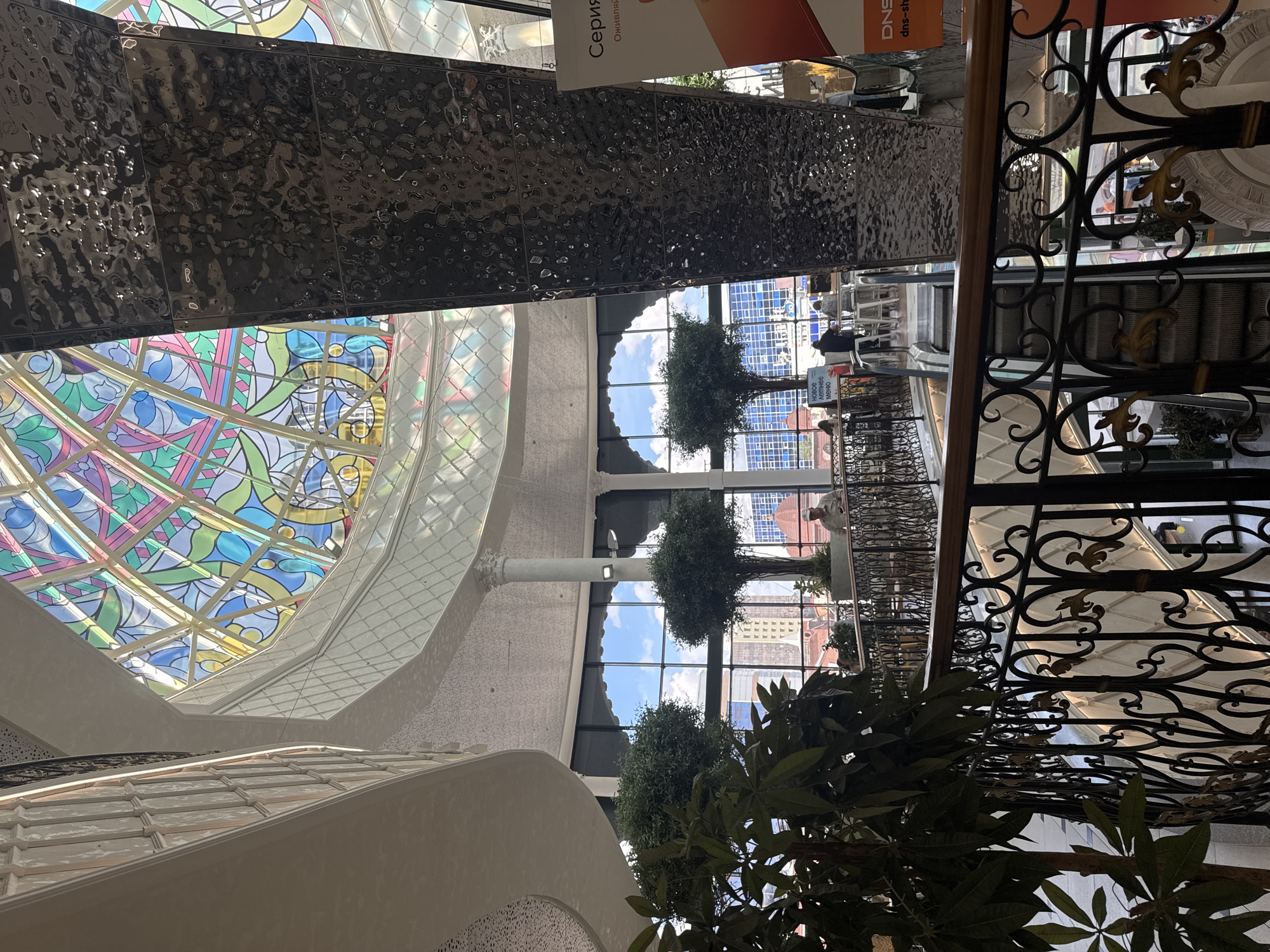The image shows the interior of a modern shopping center with a prominent stained-glass dome ceiling. The dome features a colorful, geometric pattern. In the foreground, a large pillar with a textured, metallic finish rises towards the dome. To the left, a balcony with ornate black metal railings overlooks a lower level. Several potted trees are placed on the balconies, adding greenery to the interior. Through large windows in the background, a cityscape with buildings and a cloudy sky is visible. People are seated at tables on the lower level, suggesting a cafe or dining area. Escalators are also visible, indicating multiple floors. The overall impression is of a bright, spacious, and architecturally interesting commercial space. The building appears to be the shopping center "Greenwich" in Yekaterinburg, Russia, given its distinctive stained-glass dome and architectural style.