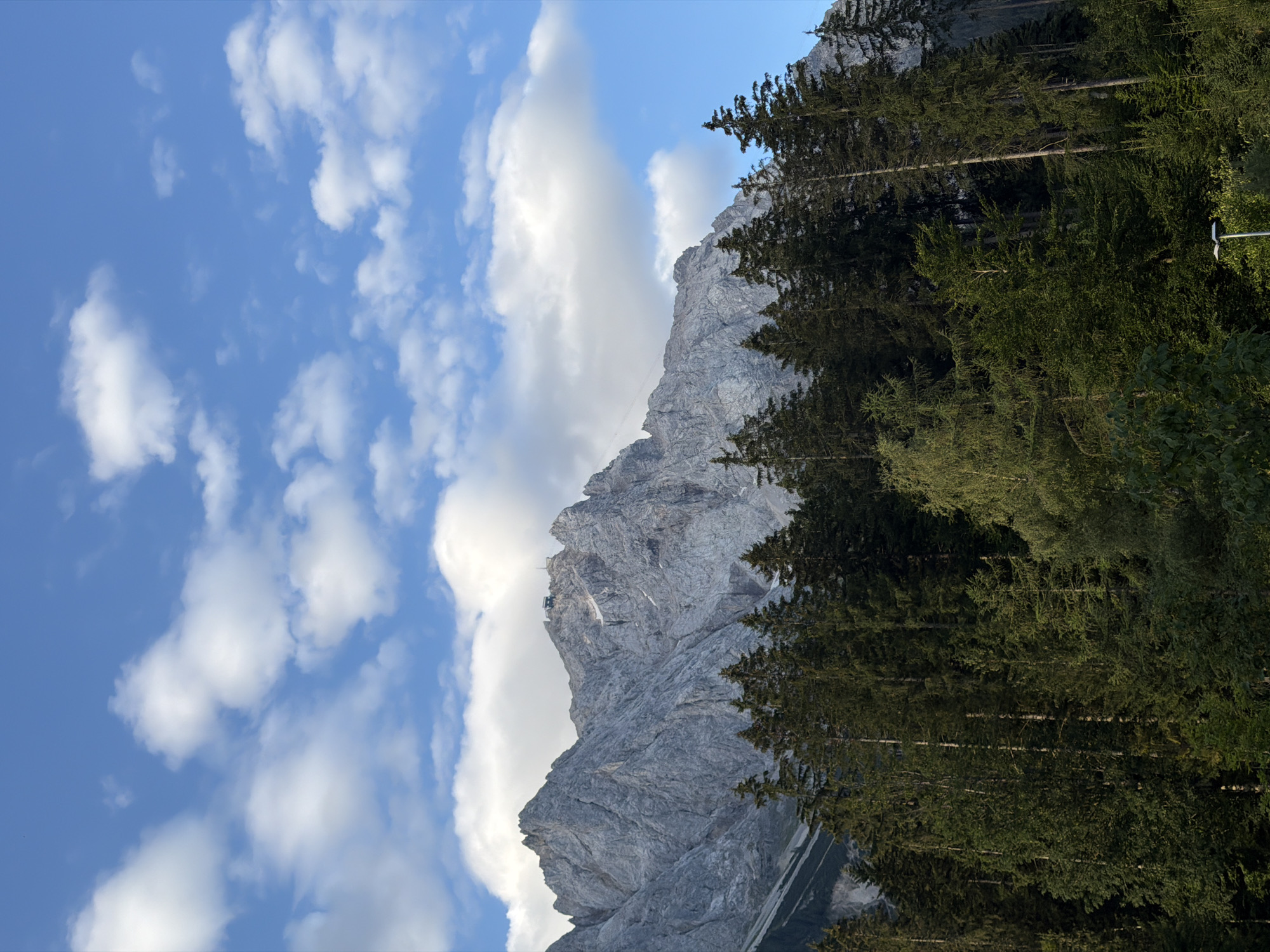 The image shows a view of the Zugspitze mountain range, the highest peak in Germany, from Grainau, Germany. The mountain is rugged and rocky, with a cable car station visible at the summit. In the foreground, a dense forest of evergreen trees covers the lower slopes. The sky above is a clear blue, dotted with white, fluffy clouds. The scene appears to be bathed in natural sunlight, suggesting it was taken during the daytime. No people are visible in the image. The overall impression is one of natural beauty and majestic scenery.
