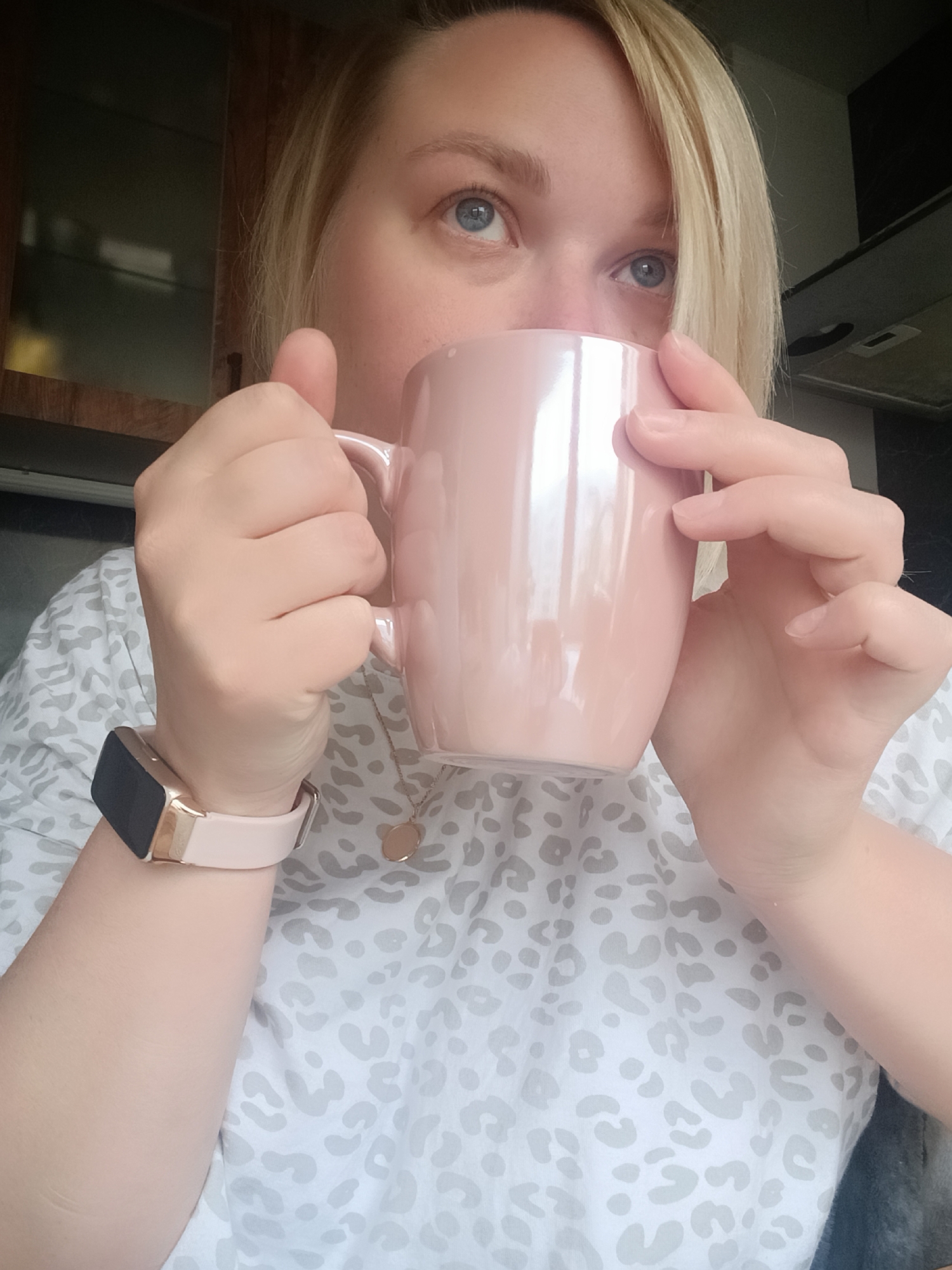 A fair-skinned person with light blonde hair holds a light pink mug, likely drinking from it. They are wearing a light-colored shirt with a gray animal print and a pink fitness tracker on their left wrist. A gold necklace is visible around their neck. The location appears to be indoors, possibly a kitchen, based on the cabinets and appliances visible in the background. It is not possible to verify that this photo was taken in Chaikovsky, Russia.