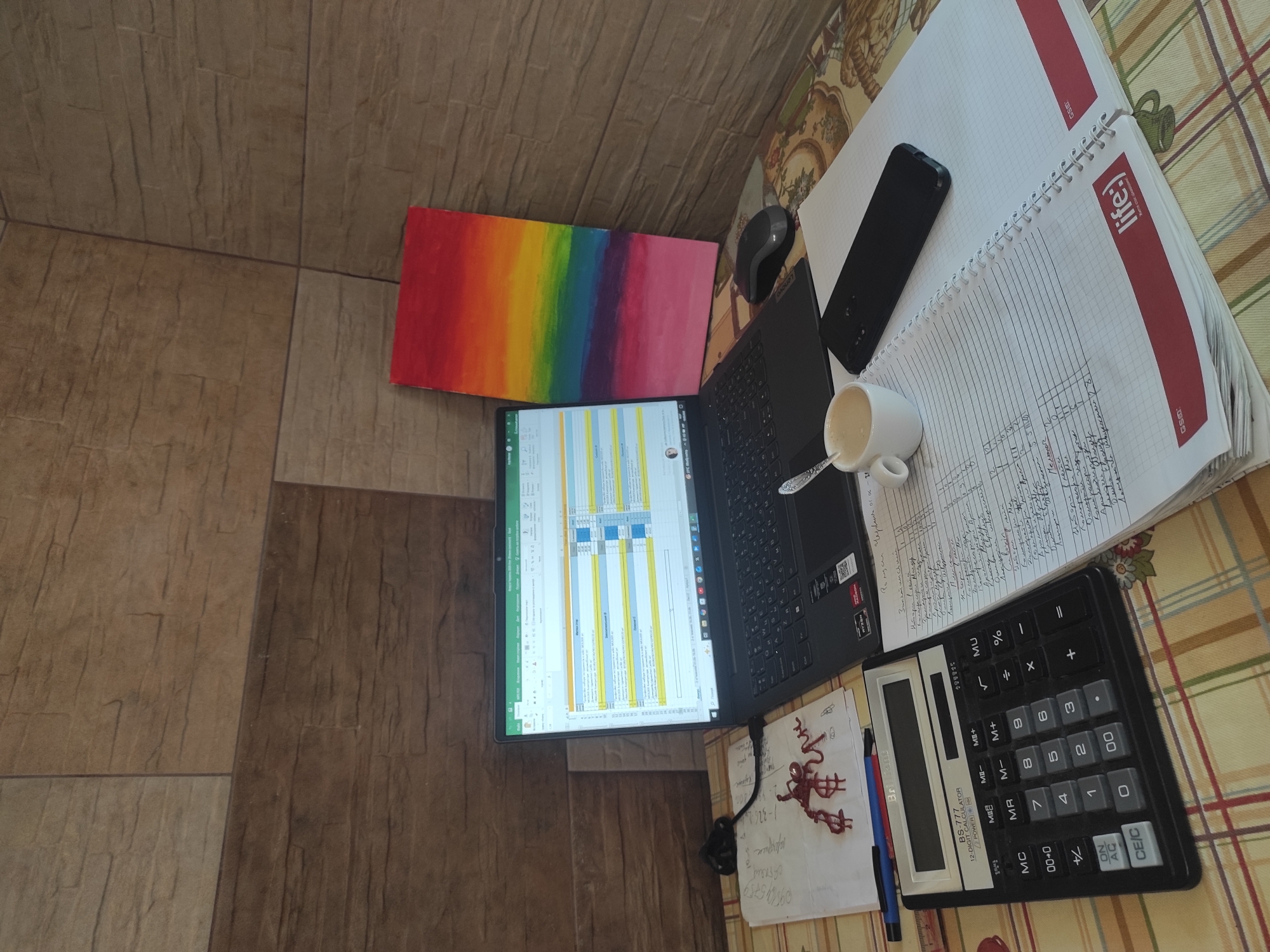 The image depicts a cluttered desk setup, likely in a home office or study, with a laptop open to a spreadsheet, a calculator, a smartphone, a notebook filled with handwritten notes, a cup of coffee, and a small, colorful painting of a rainbow. The background appears to be a textured wall, possibly made of stone or tile, with horizontal wood-like panels. The desk surface is covered with a patterned tablecloth. There is no indication of the specific location in Uzhhorod, Ukraine. The scene is static, with no visible people or ongoing actions.