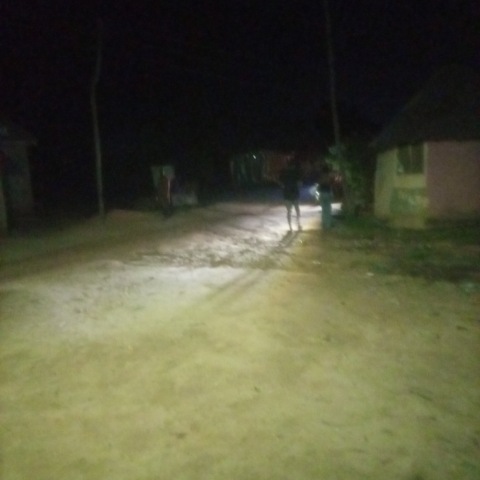 The image depicts a dimly lit outdoor scene, likely at night, with a dirt road or path in the foreground. The ground appears to be sandy or dusty, with some scattered debris or uneven patches. In the background, there are faint outlines of structures, possibly buildings, and vegetation. Two individuals are partially visible, standing on the road. One appears to be wearing dark clothing, and the other is more clearly seen as a silhouette with lighter colored pants. A tall, thin pole or tree is visible on the left side of the image. The overall lighting suggests artificial illumination from an unseen source, casting long shadows. The scene is consistent with a rural or semi-urban environment in Jalingo, Nigeria, given the dry, dusty ground and the rudimentary structures.