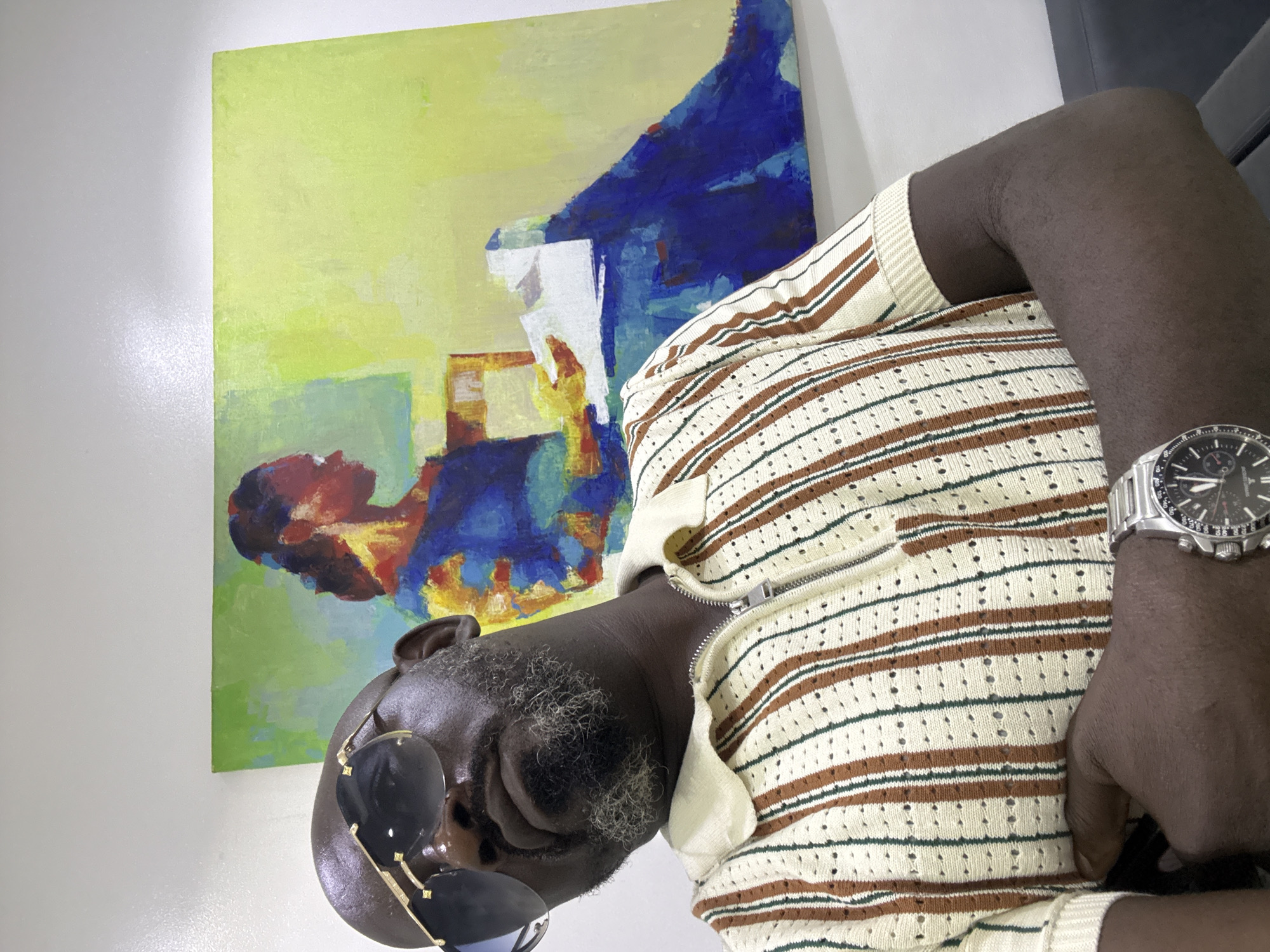 A medium shot depicts an African-American man wearing a cream and brown horizontally striped zippered top and silver wristwatch. Sunglasses are hanging on his face. Behind him is an abstract painting on a wall with green, blue, red, and yellow colors. The location is not discernable.