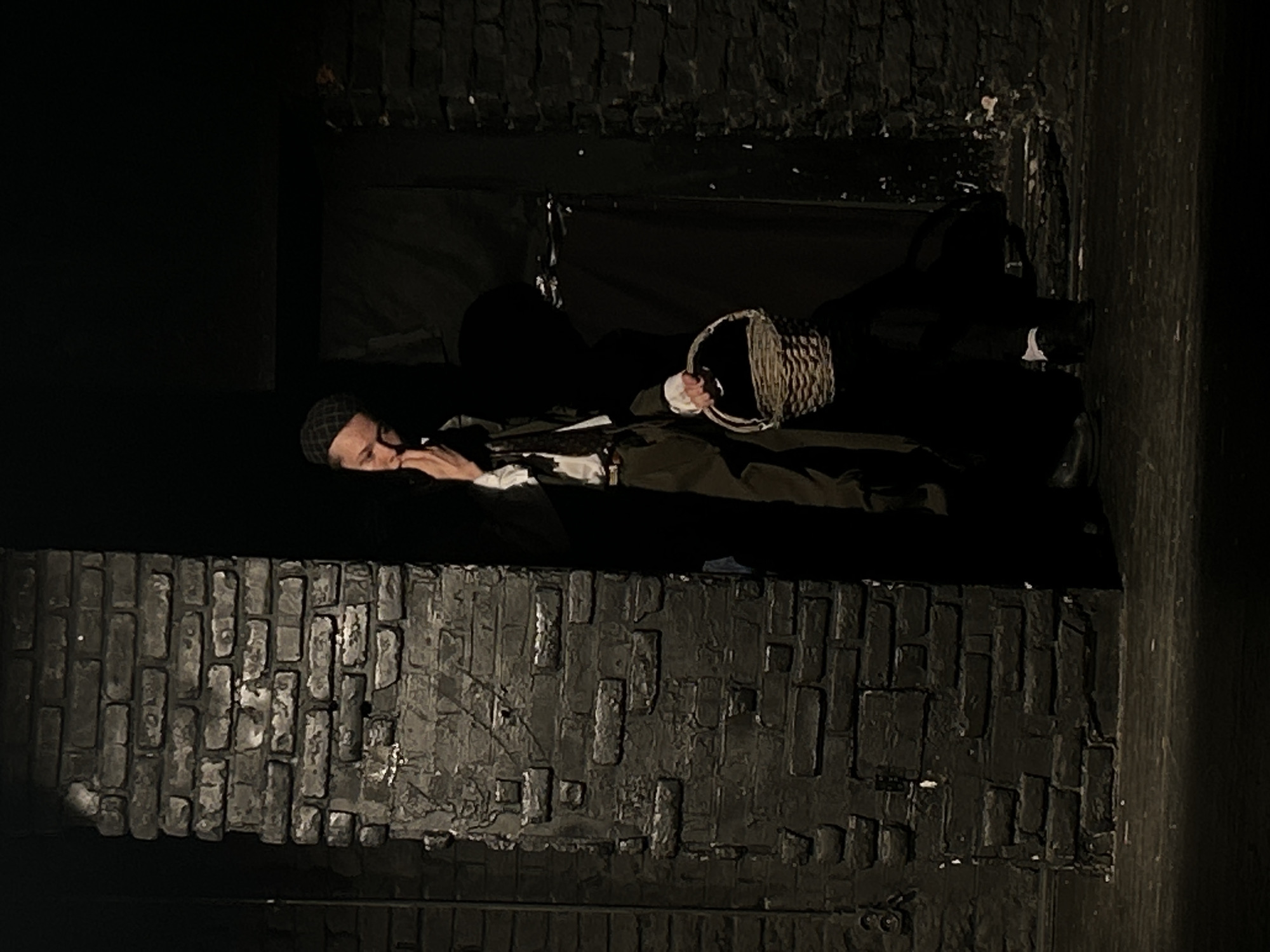 The image depicts a theatrical scene set in a dimly lit, dark environment, identified as the city of Unknown, Unknown. A single individual, appearing to be an adult, is positioned slightly left of center, emerging from behind a prominent dark brick column. The person wears a dark, possibly olive green or brown, long coat over a white shirt with white cuffs, a dark tie or ascot, and dark trousers and shoes. A dark, checkered flat cap or newsboy cap is worn on their head. The individual's left index finger is pressed against the upper lip/philtrum area, and their gaze is directed downwards and to the left, suggesting contemplation or unease. In their right hand, they hold an empty, light-brown wicker basket with a handle. The background is predominantly dark, featuring another less prominent dark brick wall on the right and a dark fabric or wall behind the person. The floor is also dark, possibly a stage, with some subtle lighter specks. Strong shadows are cast, particularly behind the individual and the brick structures, indicating a primary light source from the front-left. The overall atmosphere is dramatic and low-key.