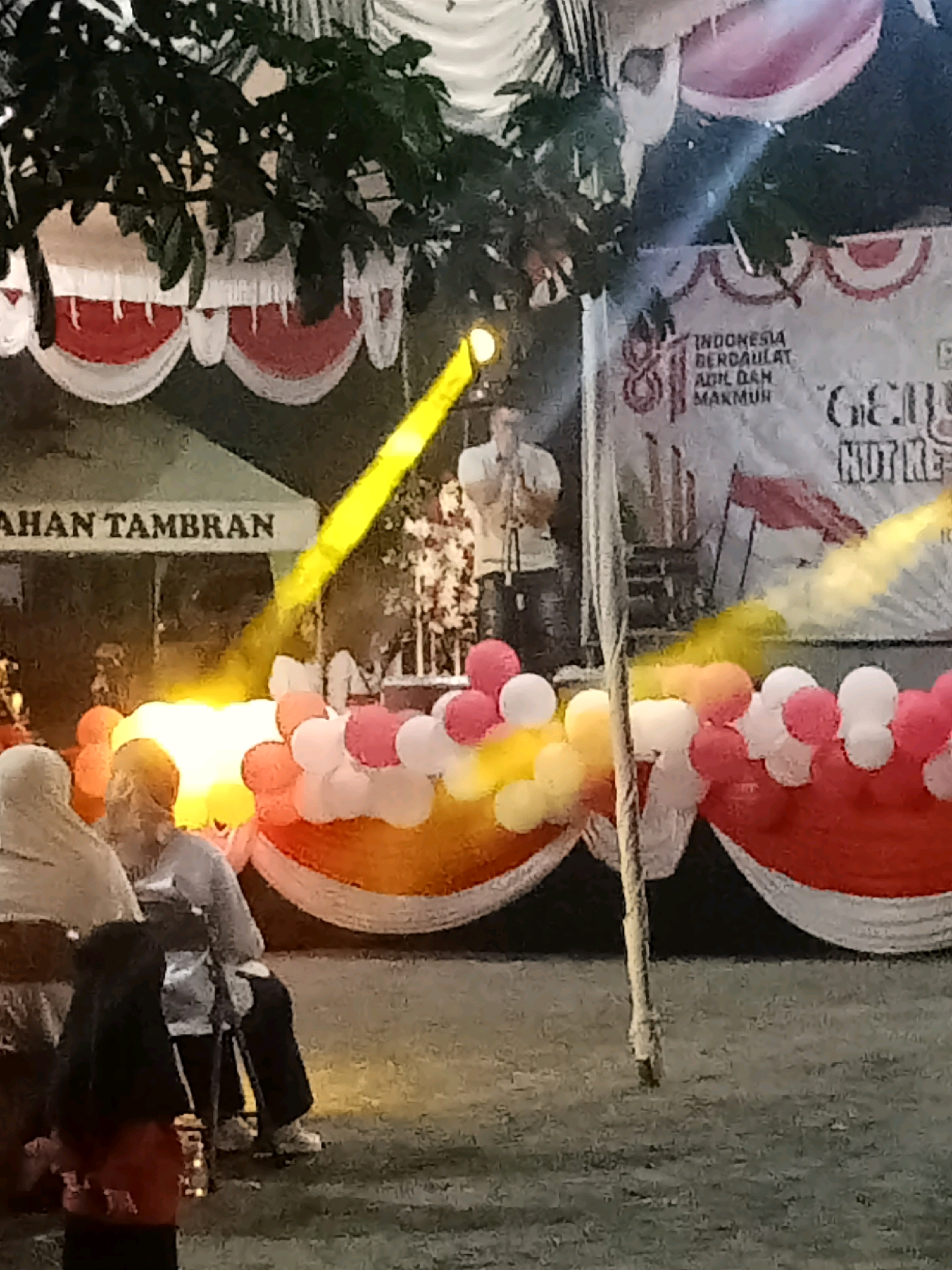 The image depicts an outdoor evening event at "Jalan Tambran," Indonesia. A stage is set up with a banner that reads "INDONESIA BERDAULAT ADIL DAN MAKMUR." A person is speaking into a microphone on stage, illuminated by a bright yellow spotlight. The stage is decorated with red and white fabric draped in a scalloped pattern, and there are clusters of pink, white, and orange balloons. Several people are visible in the foreground, some wearing headscarves and observing the event. The ground is covered in grass. The overall atmosphere appears festive.