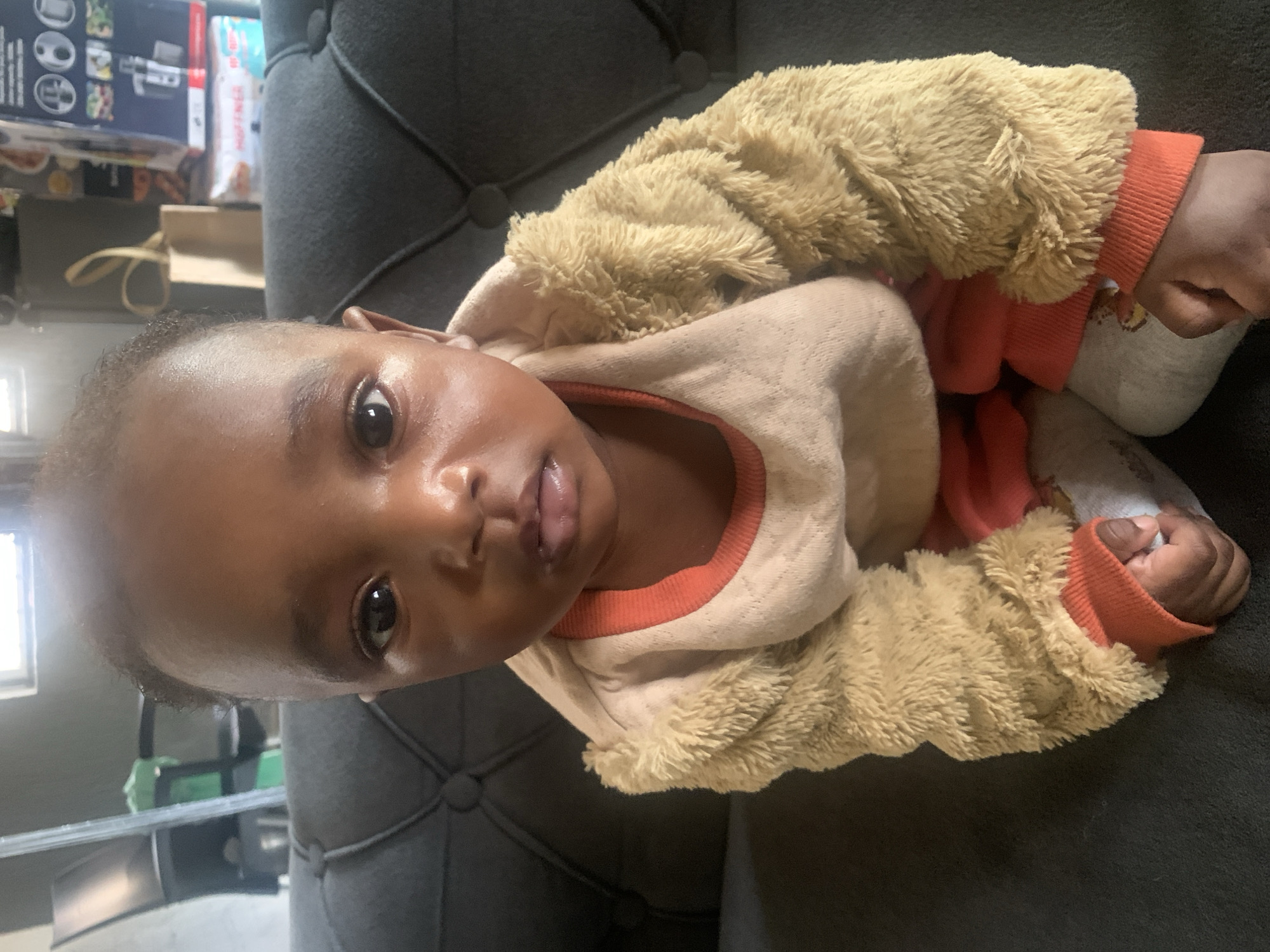 A baby lies on a dark gray tufted sofa. The baby wears a light brown fleece jacket, a beige shirt with an orange collar and trim, orange pants, and gray socks. The baby's head is turned to the left, facing the camera. The background includes a window with light entering, a green and gray object, and stacked boxes. The location is not discernible from the image.