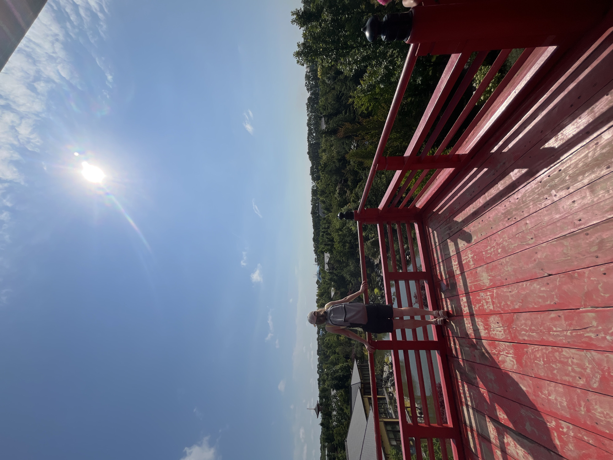The image shows a woman wearing a hat, backpack, tank top, and shorts standing on a red wooden deck overlooking a landscape. The deck has a red railing. In the background, there is a body of water, buildings with dark roofs, and lush green trees under a clear blue sky with a bright sun causing lens flare. A pagoda-like structure is visible in the distance. The scene appears to be a scenic viewpoint in a garden or park. This is not a location in Krasnodar, Russia.