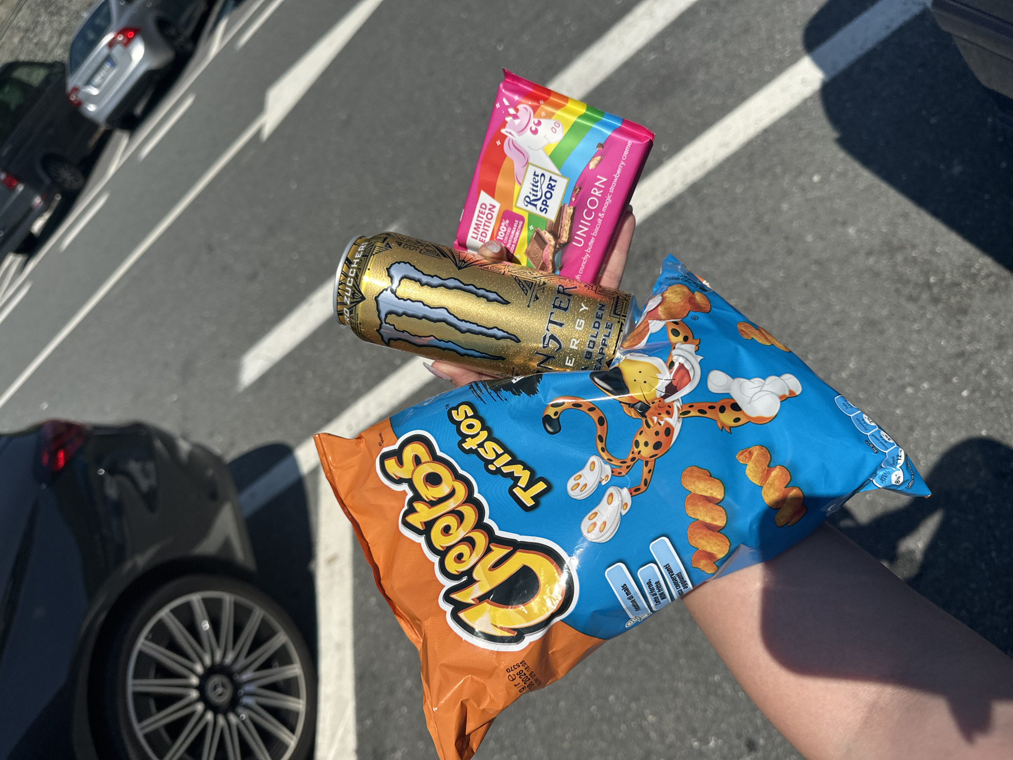 The image shows a person's hand holding a bag of Cheetos Twistos, a Monster Energy Golden Apple drink, and a Ritter Sport Unicorn chocolate bar. The hand is positioned in front of a background of parked cars and a striped asphalt surface. The image appears to be taken outdoors. Some text in the image suggests the location may be Varazze, Italy.