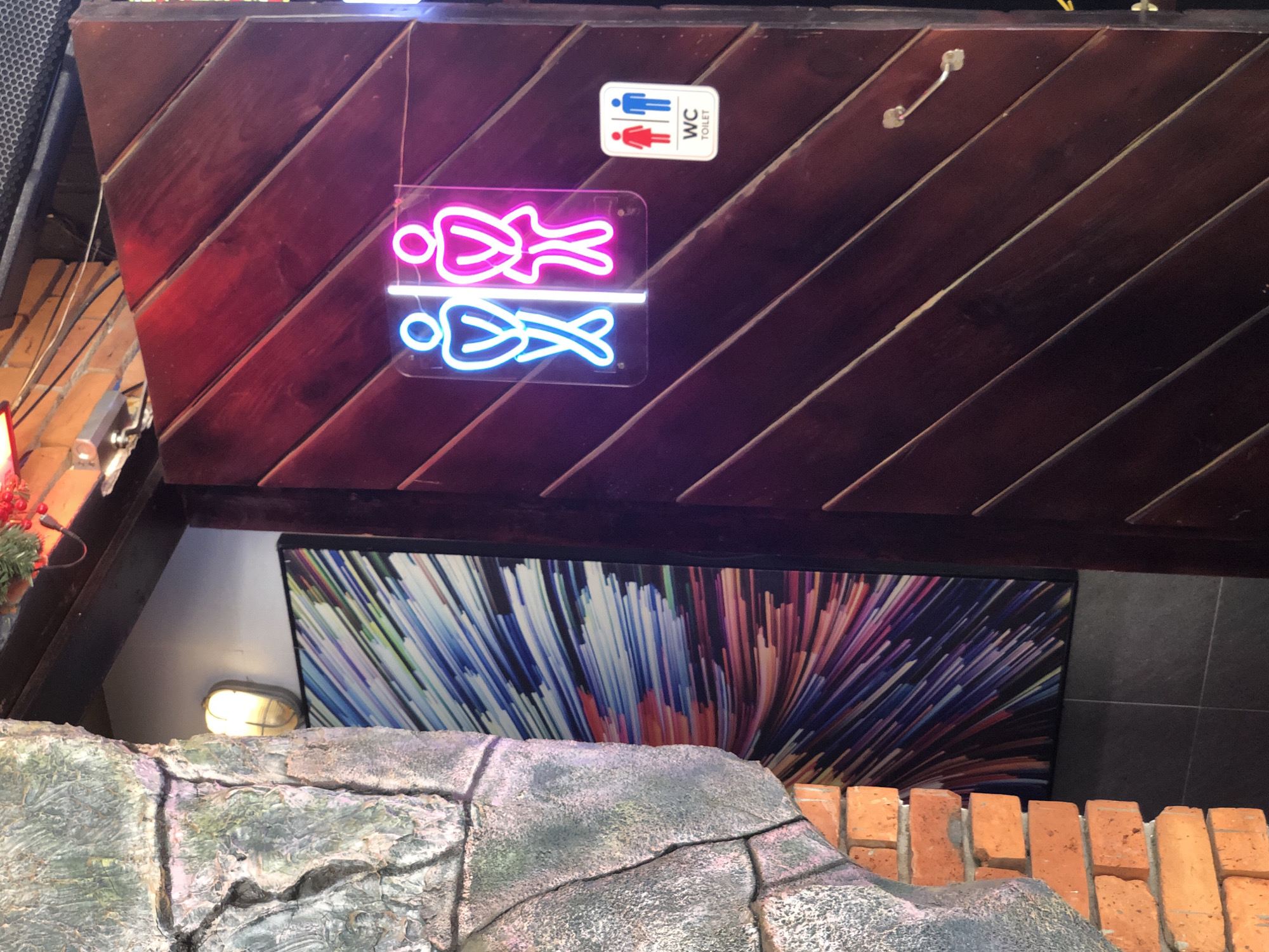 The image shows a wooden door in Đà Nẵng, Vietnam, with two illuminated signs indicating it leads to a restroom. One sign is a neon outline of male and female figures, with the male in blue and the female in pink. Below this neon sign is a smaller rectangular sign with red and blue figures representing a woman and a man, respectively, and the text "WC TOILET." The door is dark brown and made of angled wooden planks. To the left of the door, a wall is covered with rough, grey textured material, resembling stone or concrete, and beneath that, exposed orange bricks. A colorful abstract artwork is visible on the wall to the left, behind the textured material. A light fixture is mounted on the wall above and to the left of the artwork.