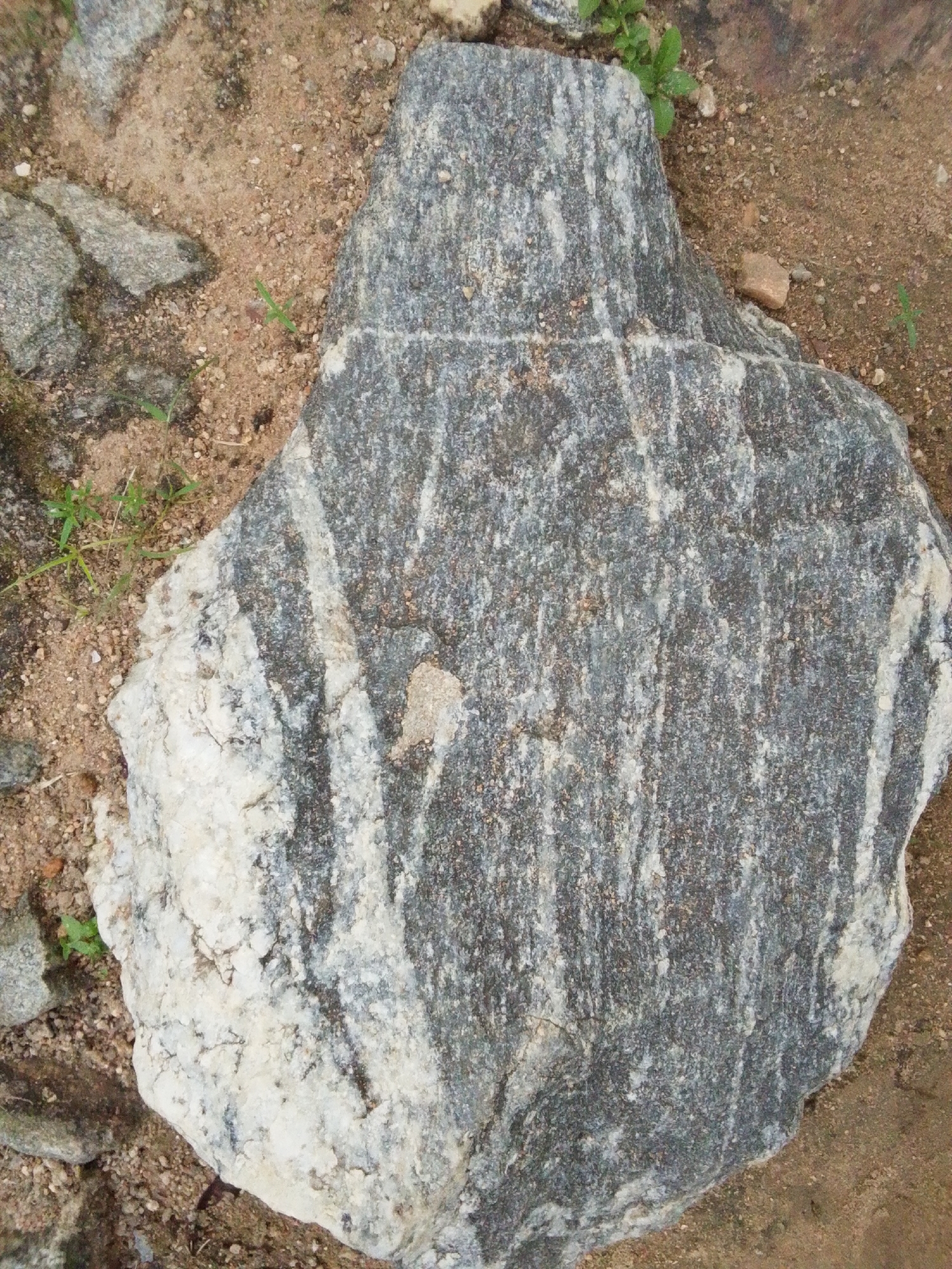 A close-up aerial view shows a large, grey, and white striped rock on sandy ground with some sparse green grass. The rock is irregular in shape, with a rough, textured surface displaying alternating bands of dark grey and lighter white and grey minerals. The ground surrounding the rock is composed of brown sand mixed with small pebbles and bits of debris. A few blades of grass are visible growing in patches of sand between the rocks. The lighting suggests an outdoor environment with natural daylight. The location is identified as Serti, Nigeria.