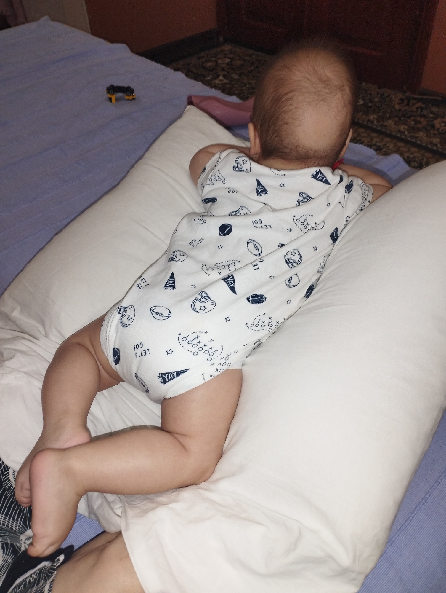 A baby is lying on their stomach on a pillow with a white onesie that has a football theme. The baby's back and legs are visible. The background shows a bed with a blue sheet and a rug on the floor. There is also a small toy car on the bed.