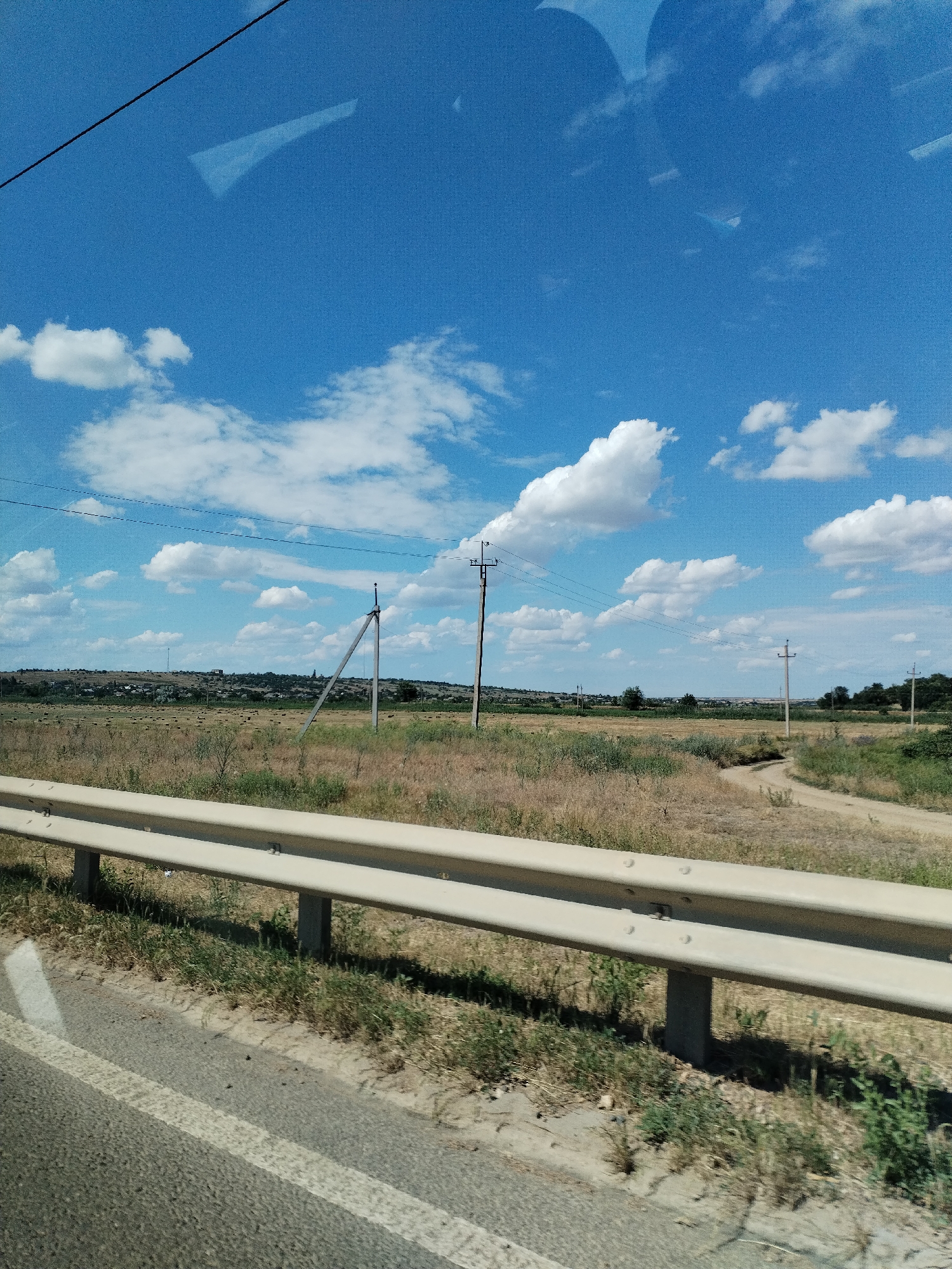 The image depicts a rural roadside scene under a bright blue sky with scattered white clouds. In the foreground, a beige metal guardrail runs along the edge of a paved road, marked with a white line. Beyond the guardrail, a dry, grassy field stretches towards a distant horizon. The field is dotted with numerous hay bales.
Several utility poles with wires are visible across the landscape, suggesting the presence of infrastructure. The landscape is gently rolling, with a small settlement of houses nestled in the distance on a low hill. A dirt road curves through the field.
The scene appears to be set during the daytime, likely late morning or early afternoon, given the bright sunlight and clear sky. The weather seems fair and warm, with the dry vegetation indicating a sunny period. The overall impression is one of a quiet, rural environment in Ukraine. There is no visible text in the image.