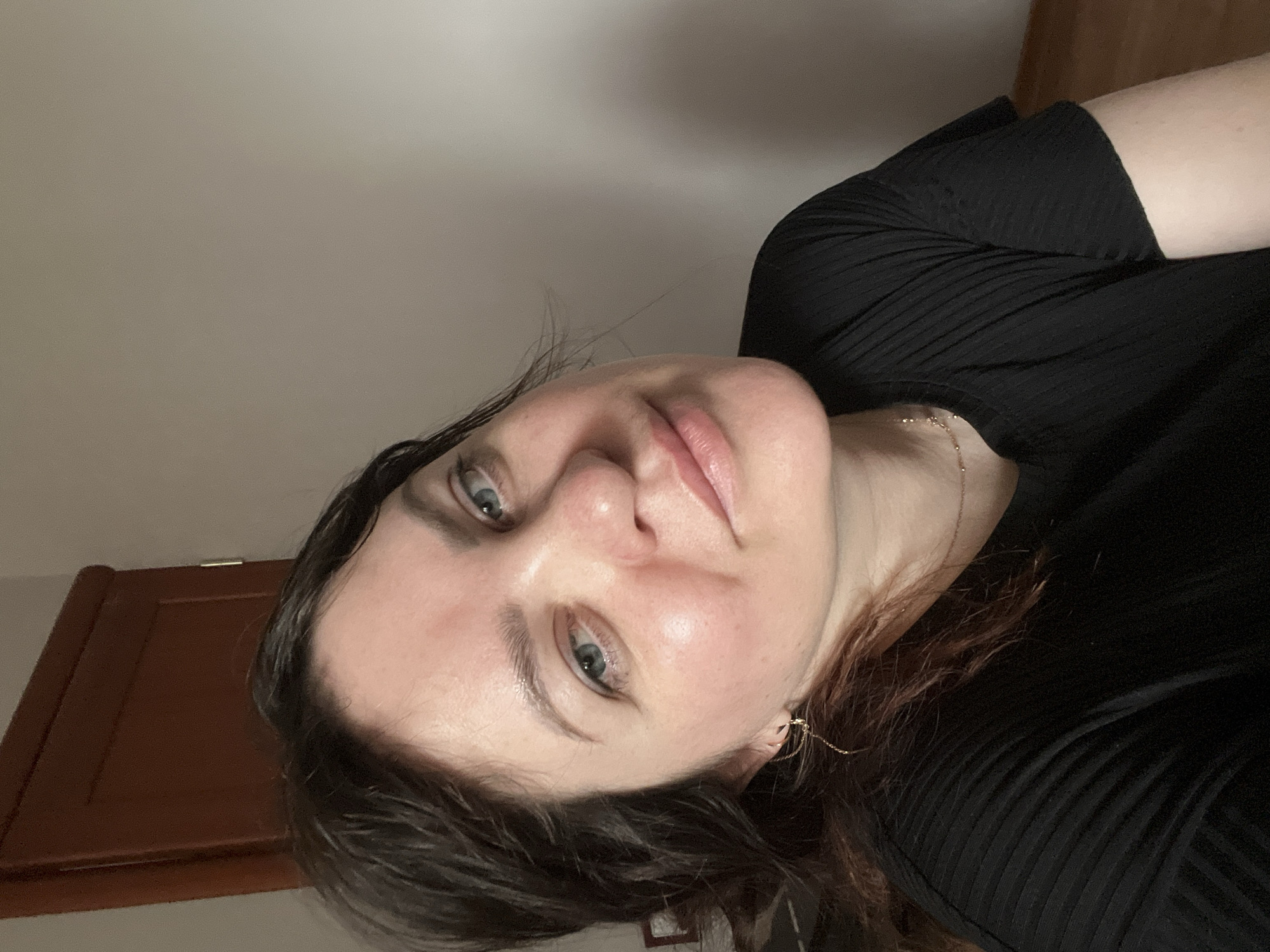 A woman with fair skin, light blue eyes, and brown hair is shown in a close-up, slightly angled shot. She is wearing a black, ribbed top. A gold chain necklace is visible around her neck. A brown cabinet is partially visible in the background. It is not possible to determine the location in Perm, Russia, from this image.