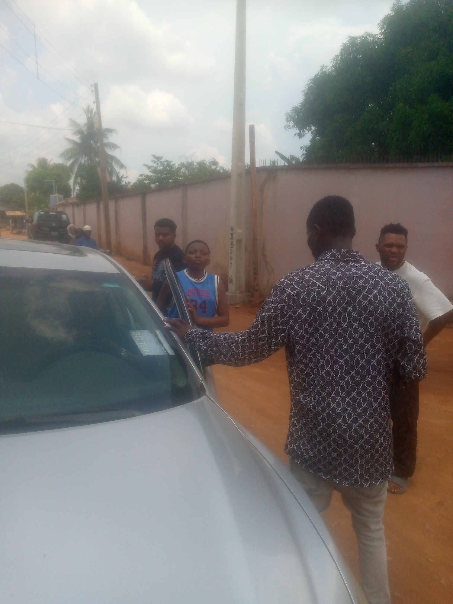 The image shows a group of people standing around a silver-colored car. There are four men and a young boy. One man is facing away from the camera, wearing a patterned shirt and jeans, and has his hand on the car door. Beside him stands a man wearing a white shirt and brown pants, with his arms crossed. Two other men stand on the other side of the car, along with a boy wearing a basketball jersey with the number "34" on it. The car has a sticker that reads "NGLOS."
In the background, there is a wall and some greenery. A utility pole is visible. Further back, there is another car and some buildings. The location appears to be a street in a residential area, city and state unknown.