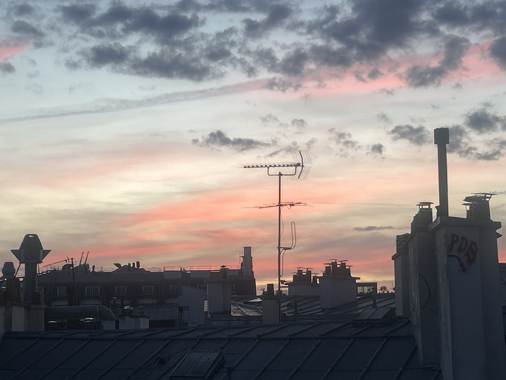 The image captures a sunset or sunrise view over a dense urban landscape, likely a European city, from an elevated perspective overlooking rooftops. The sky dominates the upper two-thirds of the frame, displaying a gradient of soft light blue, vibrant pink, and subtle orange hues, interspersed with scattered dark grey and lighter wispy clouds.
In the middle ground, numerous dark silhouettes of buildings and various rooftops are visible. Prominently centered is a tall, slender television antenna or communication aerial, with several horizontal elements, casting a slight ghosting effect, possibly from motion blur or digital artifact. Other smaller antennas and poles also punctuate the skyline. Many chimney stacks of different designs and heights are silhouetted against the colorful sky, characteristic of older city architecture. Some buildings in the mid-distance show dark outlines of windows.
On the large, rightmost chimney stack in the foreground, red graffiti spelling "PDB" is visible. The foreground features the dark, angular lines of pitched metal roofs, likely zinc or slate, with visible skylights or roof windows. The overall impression is one of a calm, twilight or dawn scene, emphasizing the distinctive rooftops and the expansive sky. There are no people visible, and no specific activity is taking place beyond the natural progression of daybreak or dusk.