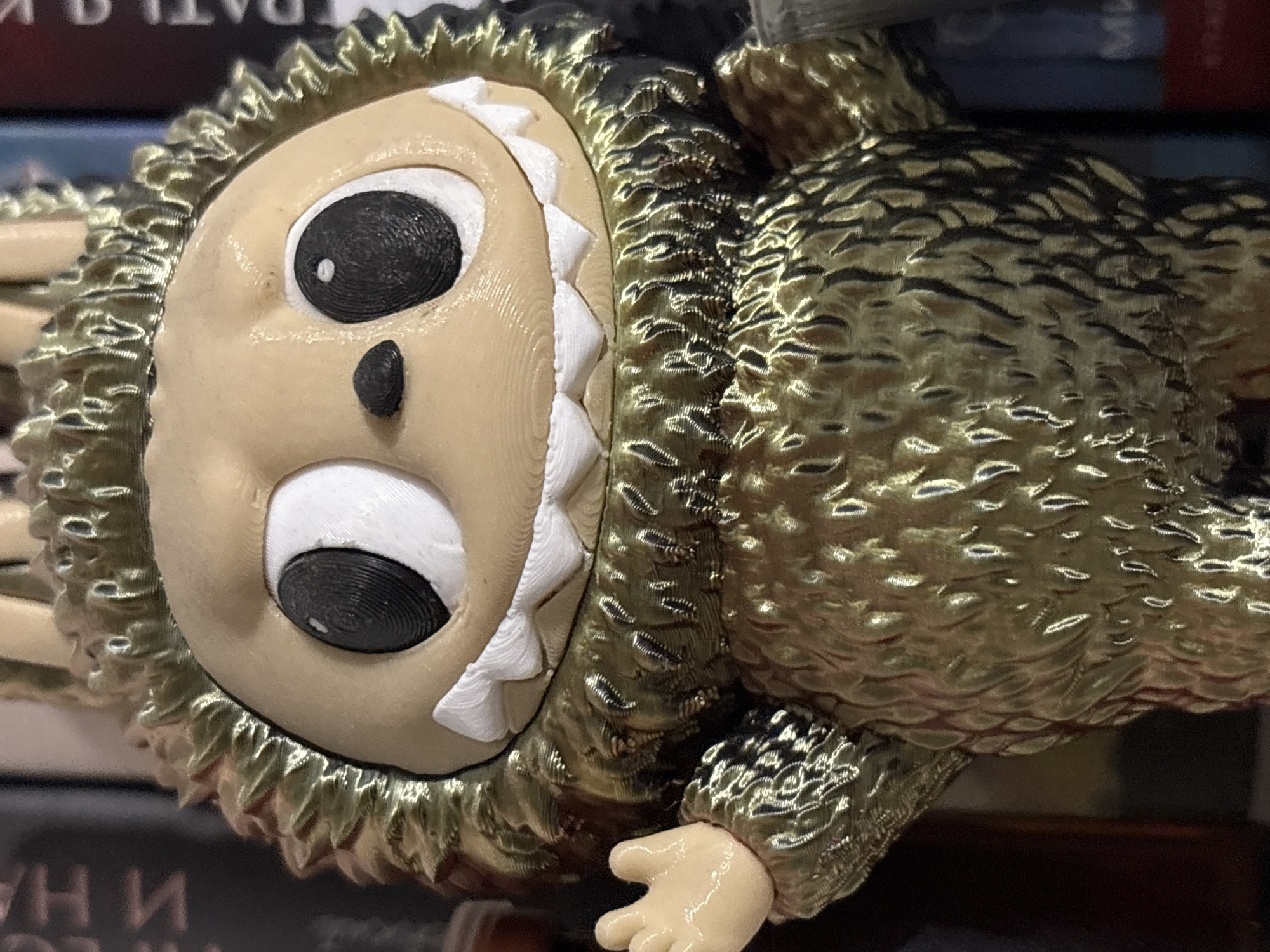 A close-up shot shows a 3D-printed figure with a tan face, large black eyes, and a small black nose. A set of white, triangular teeth lines the edge of the face. The figure has a golden, textured body resembling feathers or scales. A small, tan hand extends from the body. The figure sits atop books, with text partially visible on the book covers beneath. The location and city are not discernible from the image.
