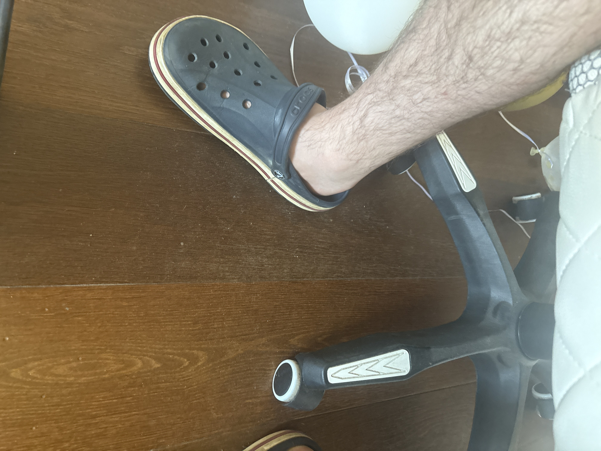 The image shows a close-up view of a person's right leg and foot wearing a dark blue Croc shoe. The leg is hairy and appears to be bare from the knee down. The Croc shoe has the characteristic holes on the front and a striped sole with red and cream-colored bands. The shoe is positioned on a wooden floor. In the background, a black office chair with a white padded seat is visible, along with parts of its base and wheels. A white object, possibly a balloon, is also partially visible to the left of the leg. The lighting suggests an indoor setting. The scene is observed from a slightly elevated angle, looking down towards the floor. There is no visible indication of the city of Orekhovka, Russia.