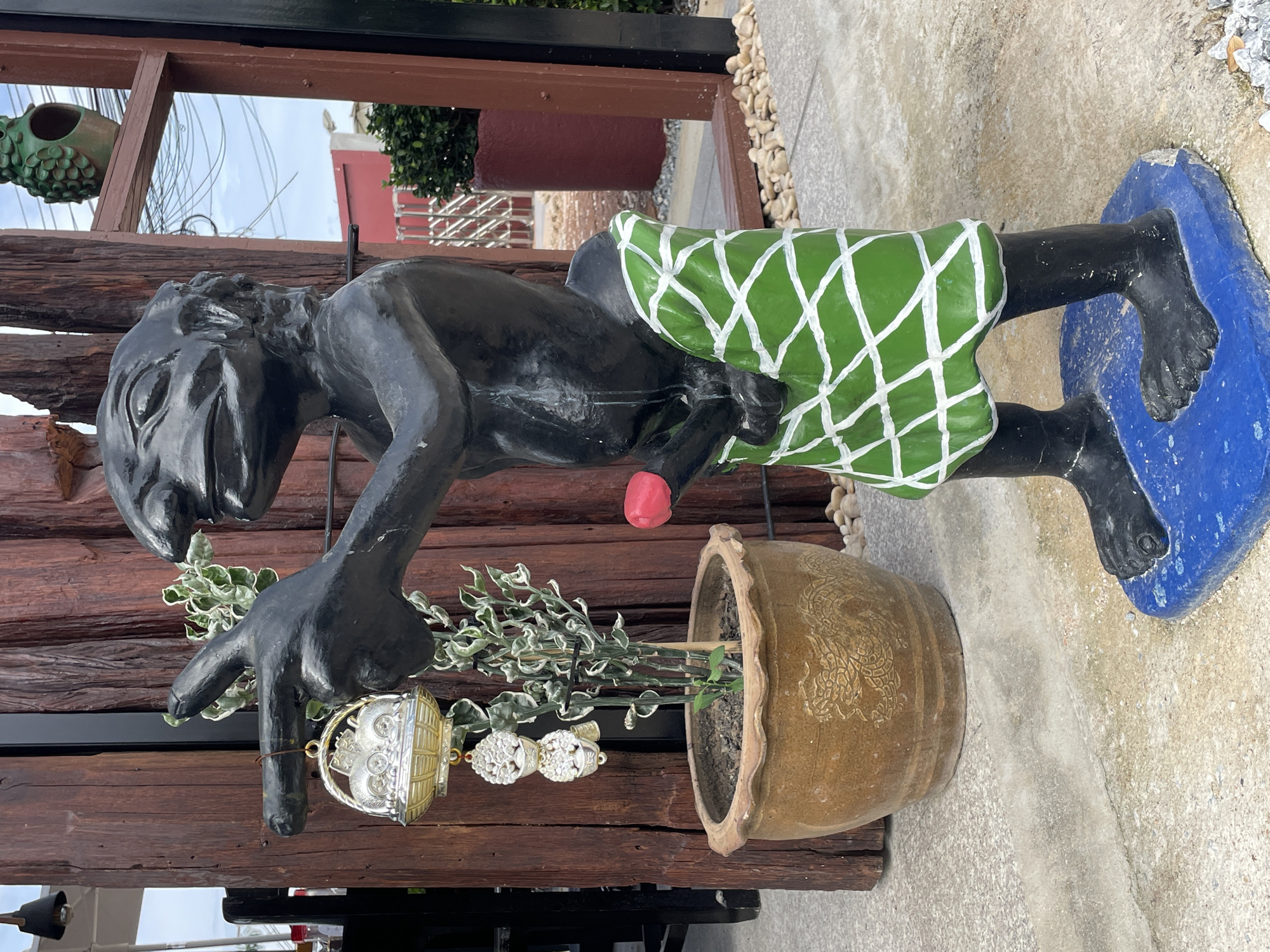 In Nong Prue, Thailand, a dark-colored, statue of a creature with exaggerated facial features stands on a blue base. The statue is partially covered by a green loincloth adorned with white crisscrossing lines. A red, phallic-shaped object is attached to the statue's midsection. To the left of the statue, a potted plant with green and white variegated leaves sits in a brown ceramic pot with dragon motifs. A silver and gold decorative hanging ornament with three owl-like figures is suspended from the statue's outstretched arm. The background shows a wooden structure, a reflection in a window, and some vegetation.