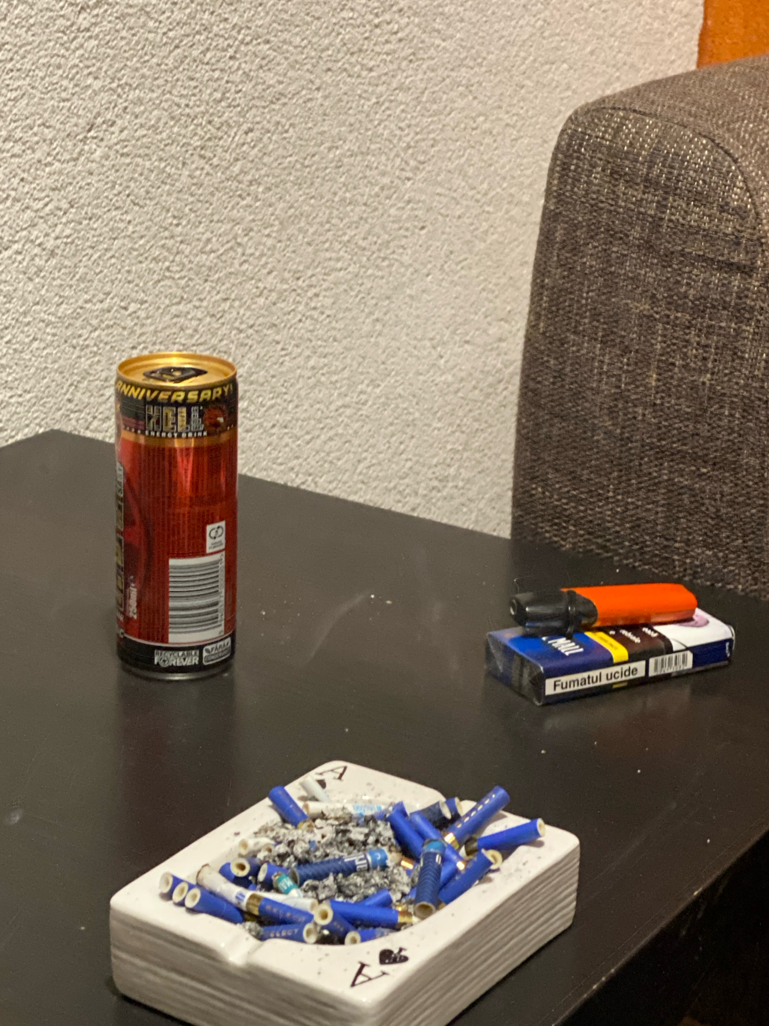 The image depicts a close-up, eye-level view of a dark, flat surface, likely a table, with several objects arranged on it. The background consists of a textured, light-colored wall and a portion of a brown, textured piece of furniture.
In the foreground, a white ceramic ashtray is filled with numerous cigarette butts and ash. The cigarette butts are of various types, with some featuring blue filter tips, while others appear to have white or light-colored filters. The ashtray itself has a distinctive pattern of playing card symbols (hearts and possibly spades or clubs) embossed on its sides.
To the left of the ashtray stands a red and silver aluminum can of "HELL ANNIVERSARY ENERGY DRINK." The can has a gold lid and features prominent branding and nutritional information.
To the right of the energy drink can, and behind the ashtray, is a blue and white pack of cigarettes. Resting on top of the cigarette pack is an orange and black electronic cigarette or vape device. The cigarette pack displays text in what appears to be Romanian, including the warning "Fumatul ucide" (Smoking kills).
The lighting in the scene is dim, casting subtle shadows and highlighting the textures of the objects. The overall impression is one of a casual, perhaps solitary, setting. The image does not provide any explicit indication of the specific location within Hodoș, Romania, beyond the general context of an indoor environment. There are no people visible in the image, nor any actions or interactions taking place. The scene is static and focused on the arrangement of the depicted items.