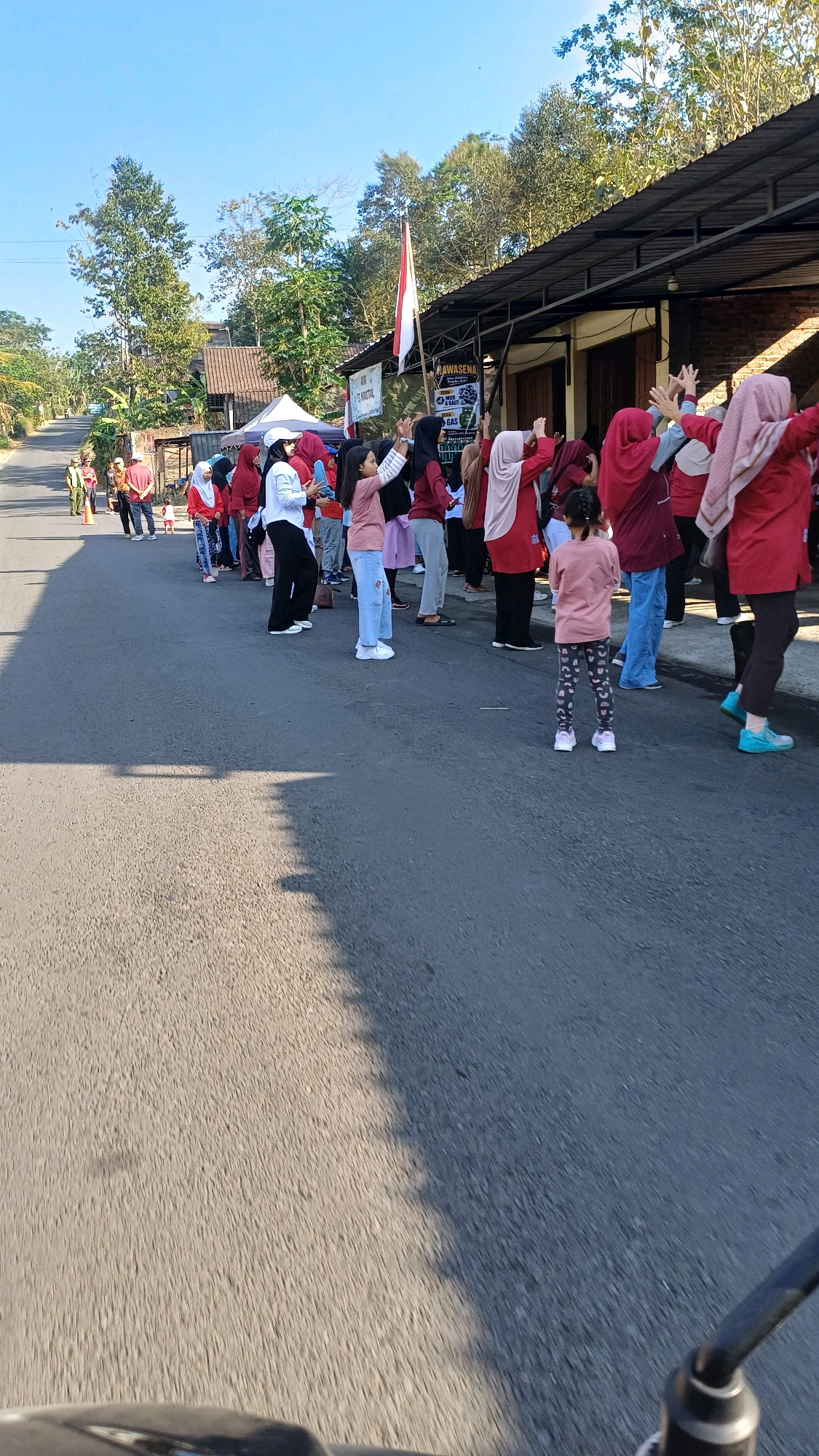 In the city of Lungge, Indonesia, a group of individuals, predominantly women and children, are gathered on a paved road, participating in an outdoor activity, possibly a form of exercise or a community event. The group is lined up, facing towards a building with a dark, sloped roof structure, possibly a shop or community hall. A red and white Indonesian flag is visible, attached to a pole extending from the building. The participants are dressed in casual clothing, with many wearing red or pink tops. Some individuals are raising their arms in unison, suggesting a coordinated movement. The road appears to be a rural or semi-urban street, with trees and houses visible in the background. The weather is clear and sunny, with a bright blue sky. A shadow is cast across the road, indicating the time of day. The image is captured from a low angle, possibly from a vehicle, with the foreground showing a portion of a handlebars and a wheel.