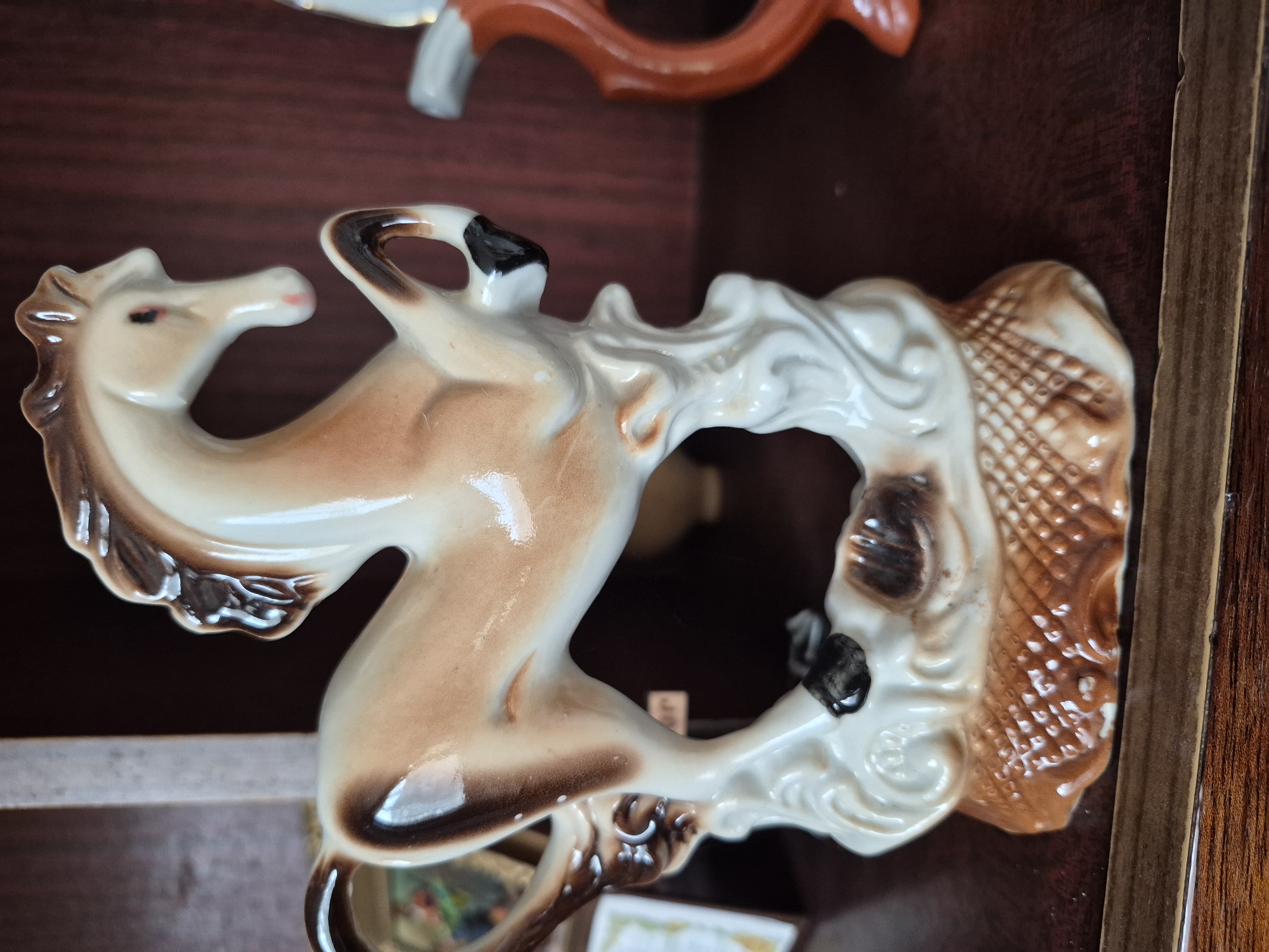 A ceramic figurine of a horse is positioned on a wooden shelf. The horse is a light tan with darker brown markings on its mane, tail, and legs. It stands on a base that resembles a textured woven pattern. The background is dark and appears to be a shelf or cabinet. To the right of the horse, another figurine, partially visible, is orange and has a curved shape, possibly part of a set or another decorative item. The scene is indoor and likely domestic. The location of Vanadzor, Armenia is not ascertainable from the image.