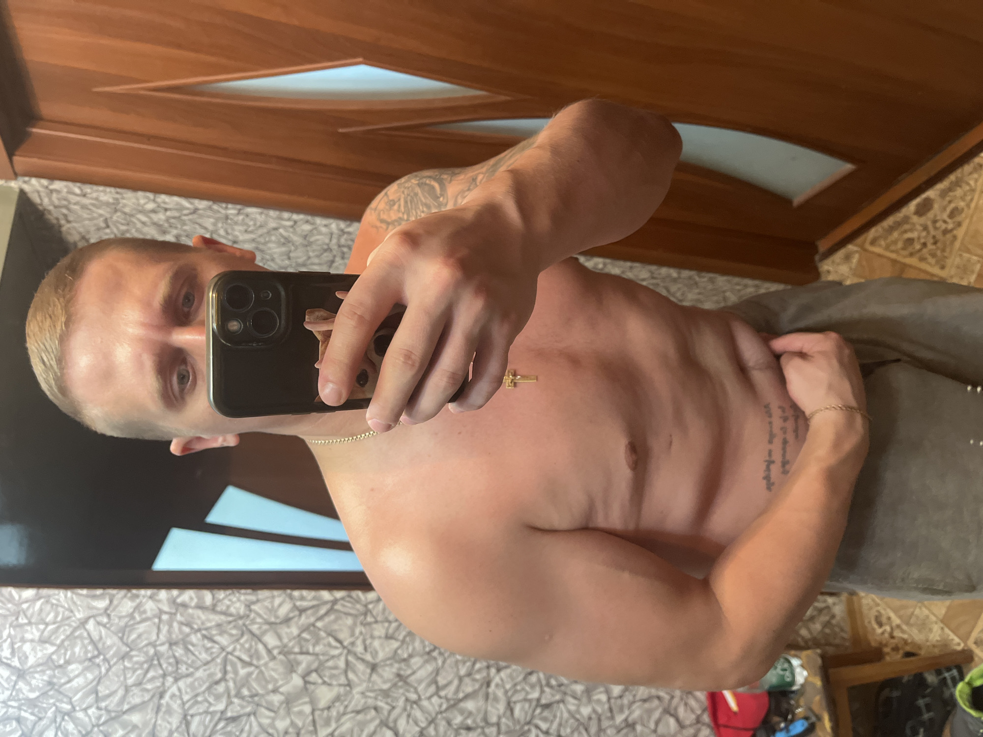 The image is a mirror selfie taken indoors by a shirtless man with short blond hair, light blue eyes, and a tattoo on his right arm. He is wearing grey sweatpants and a gold chain with a cross pendant. The background shows a textured wall, a wooden door with glass inserts, and a tiled floor. A small table with various items, including a bottle and a red object, is visible on the left side of the frame. The scene appears to be a residential interior, possibly a bathroom or bedroom, and there is no indication of the image being taken in Minsk, Belarus.
