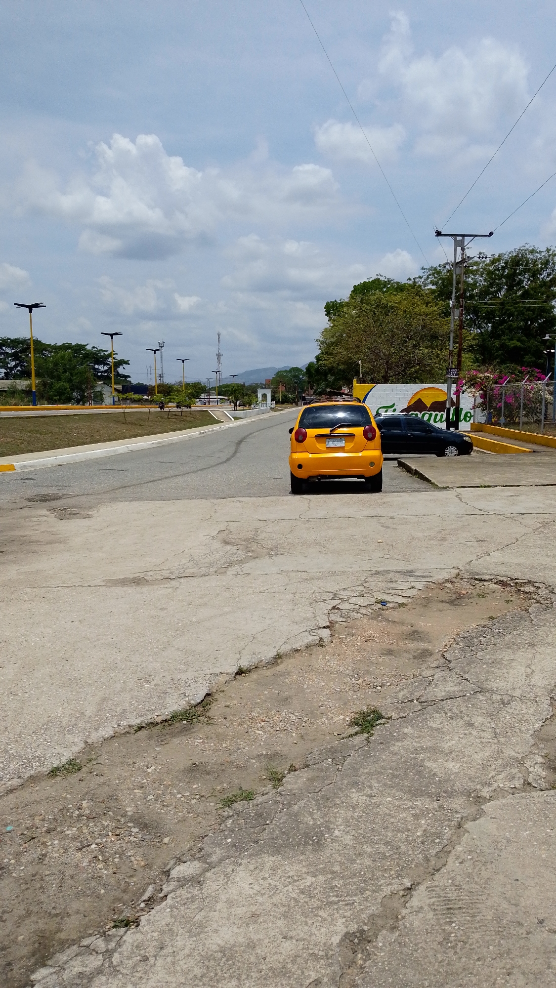The image captures a daytime scene in Tinaquillo, Venezuela, under a partly cloudy sky. The main subject is a bright yellow compact car, likely a Chevrolet Spark, positioned in the middle ground with its rear facing the viewer. Its license plate, "AF-005PV," is visible. The car is on a paved road that curves slightly to the left, flanked by a grassy median on the left.
In the immediate foreground, the ground is a mix of cracked and uneven concrete and dirt with sparse patches of grass, indicating wear. Further down the road on the right, a dark-colored sedan is parked beside a colorful wall. This wall features a vibrant mural with yellow and green colors, and the word "Tinaquillo" is clearly visible.
The scene includes several streetlights with distinctive, modern designs lining the road and the median. Utility poles with overhead wires are also present on the right side of the road. Lush green trees and vegetation frame the scene, with some flowering bushes adding a touch of color near the mural. In the distance, mountains are visible under the expansive sky, and a white archway marks a potential entrance or landmark further down the road. The overall atmosphere suggests a quiet, semi-urban or rural setting. No people are visible, and there is no overt activity taking place beyond the parked vehicles.