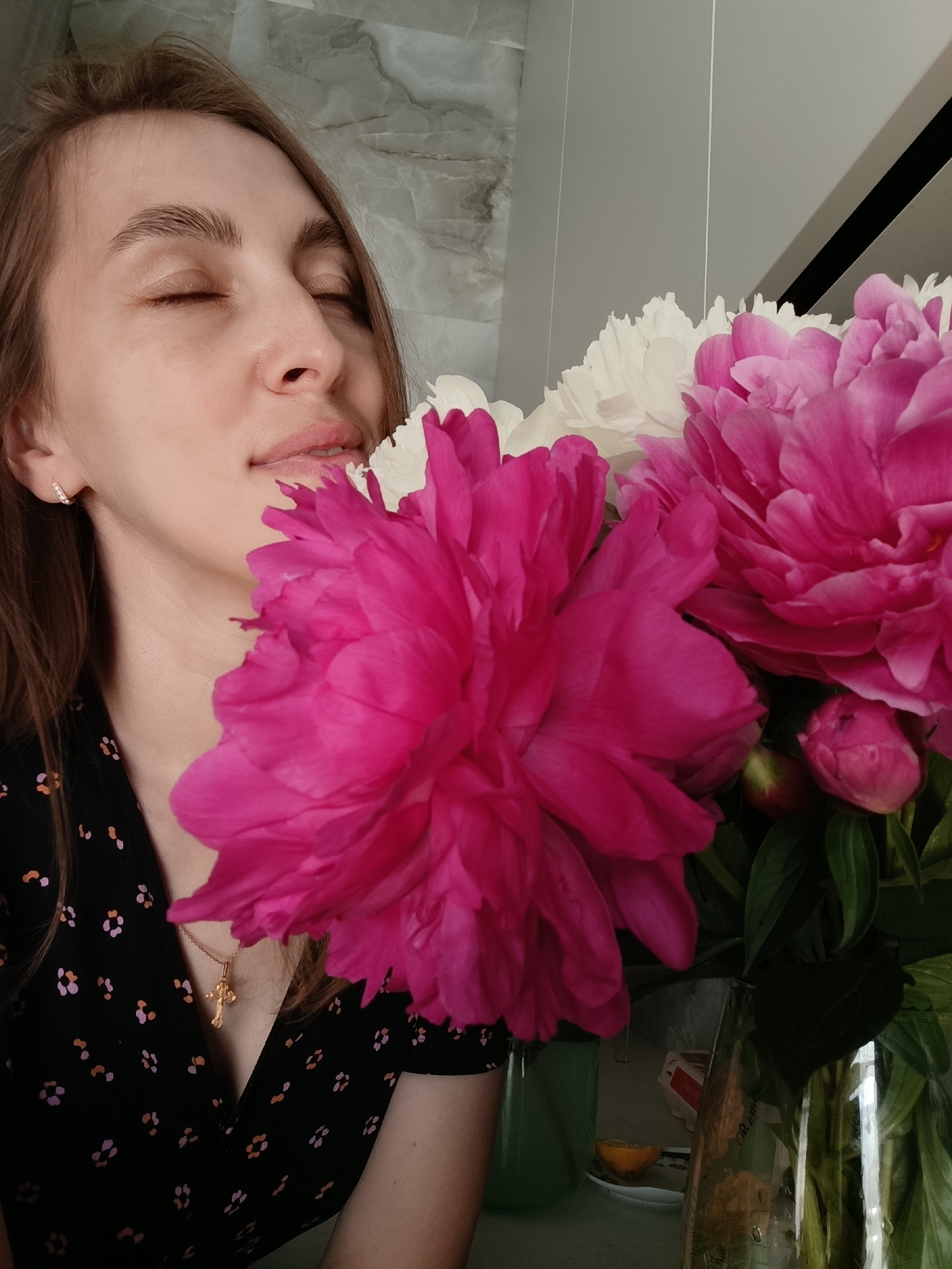 A medium shot captures a woman with closed eyes, appearing to inhale the fragrance of a bouquet of peonies. She is positioned on the left side of the frame, her head tilted slightly upwards. Her brown hair is parted and falls around her face. She wears a black dress with a subtle floral pattern and a gold cross pendant. The bouquet, in the foreground, features large, vibrant pink and white peonies. The background includes a wall with a marble-like texture and a light grey, possibly metallic, surface. To the right of the bouquet, a glass vase holds the flowers and some greenery. The scene appears to be indoors, possibly in a domestic setting within the city of Chaikovsky, Russia.