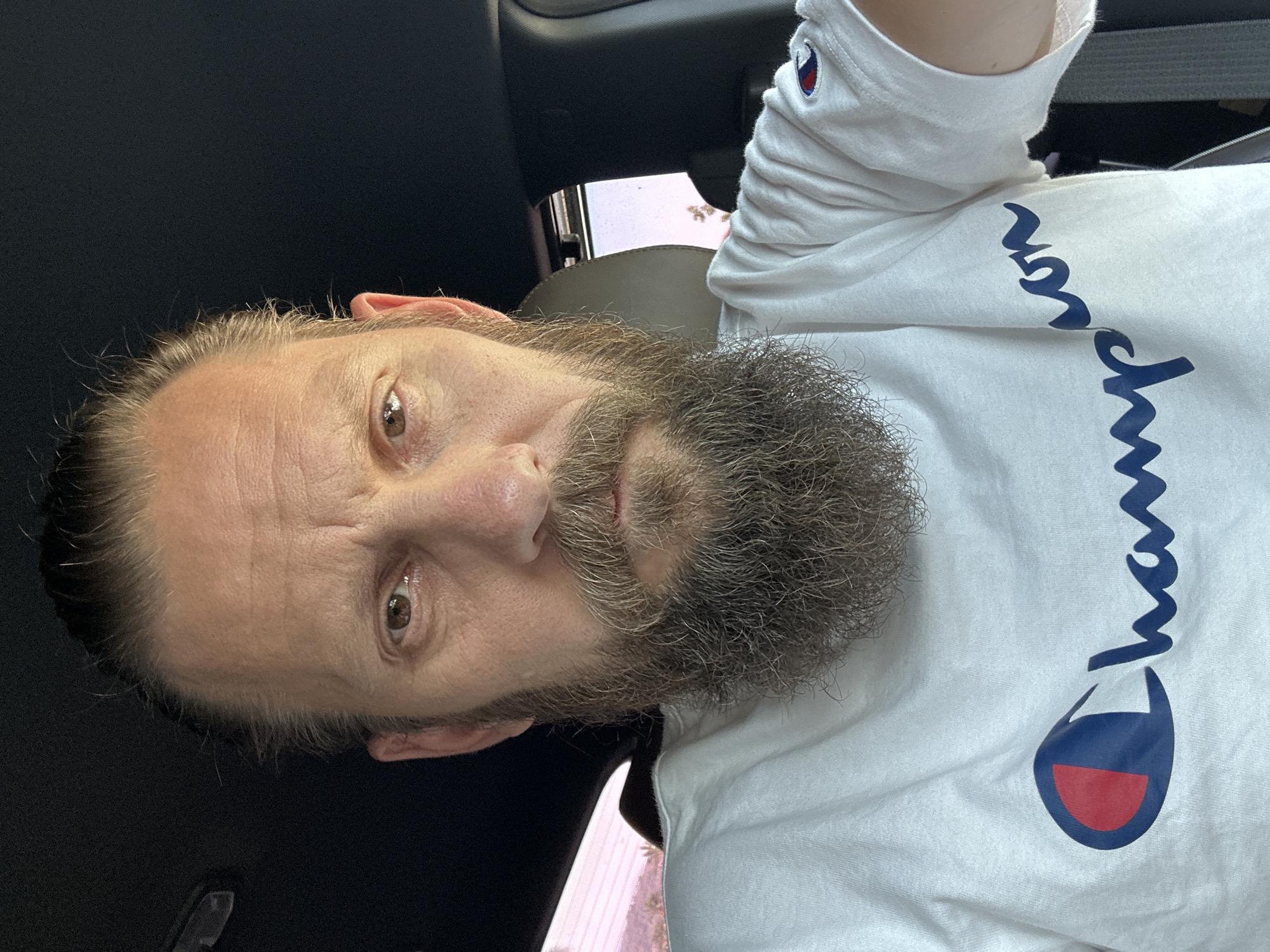 A man with a full beard and a bun hairstyle is looking directly at the camera, centered in the frame. He is wearing a white Champion t-shirt with the logo visible on the chest and sleeve. The man appears to be inside a vehicle, suggested by the dark interior of the car and a glimpse of a seat headrest. The background is blurred, showing a hint of sunlight and possibly some vegetation through a window. The overall lighting is bright, and the image is a close-up of the man's face and upper torso. The location is not specified as Fethiye, Turkey.