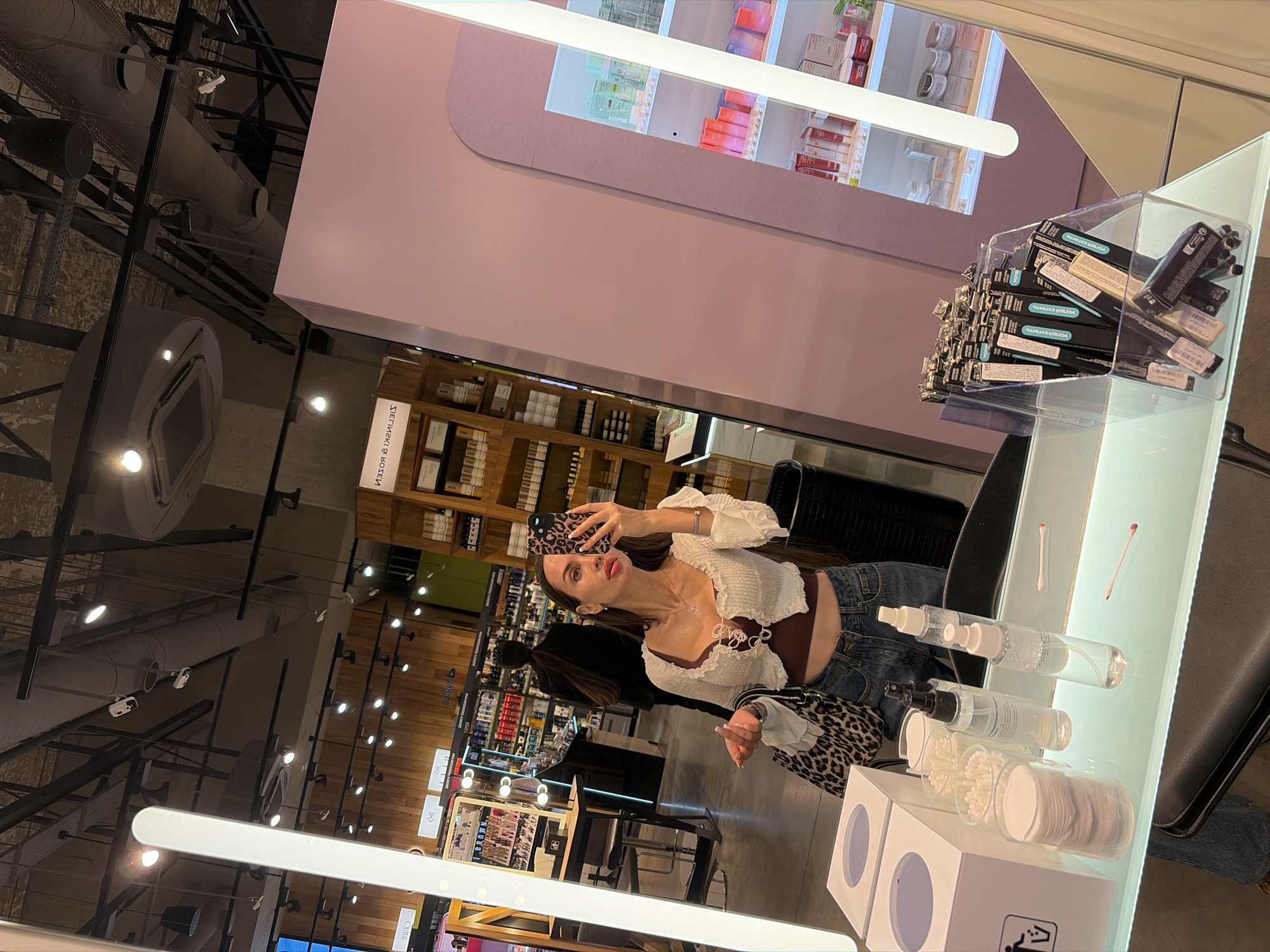 A woman is taking a mirror selfie in a brightly lit cosmetics store. She is wearing a white cropped top with a dark red camisole underneath and blue jeans. She has long dark hair and is wearing makeup, including bright red lipstick. In her right hand, she is holding a phone with a leopard-print case, and in her left hand, she is holding a leopard-print bag.
The store appears to be a modern retail space with shelves stocked with beauty products. There are large mirrors and display cases. In the background, another person can be seen walking away. The overall lighting is bright and focused on the product displays and the woman. The environment suggests a clean and organized retail setting. The store is located in Saint Petersburg, Russia.