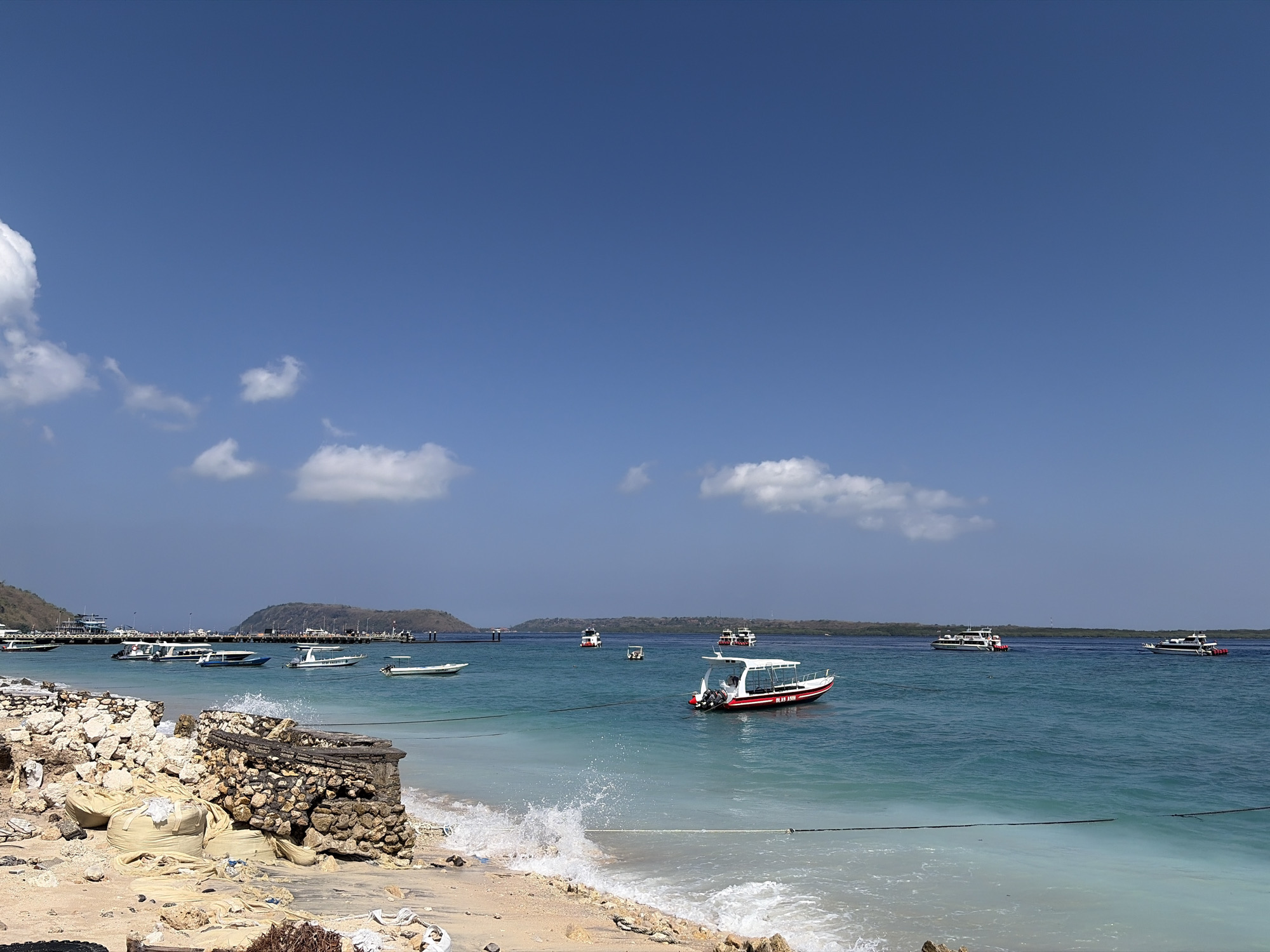 The image shows a coastline in Nusa Penida, Indonesia. The foreground features a sandy beach with scattered debris, including large sandbags and rocks. A partially constructed stone barrier is visible. The turquoise water of the ocean is clear and calm, with small waves breaking on the shore. Several boats are anchored or moving in the water, including a red and white speedboat in the mid-ground and several larger passenger boats and smaller speedboats further out. In the background, a long pier extends into the water, with more boats docked. Hills with sparse vegetation form the distant coastline. The sky is a clear, bright blue with a few scattered white clouds. The scene depicts a typical day at a coastal area, likely a port or tourist destination, in Nusa Penida.