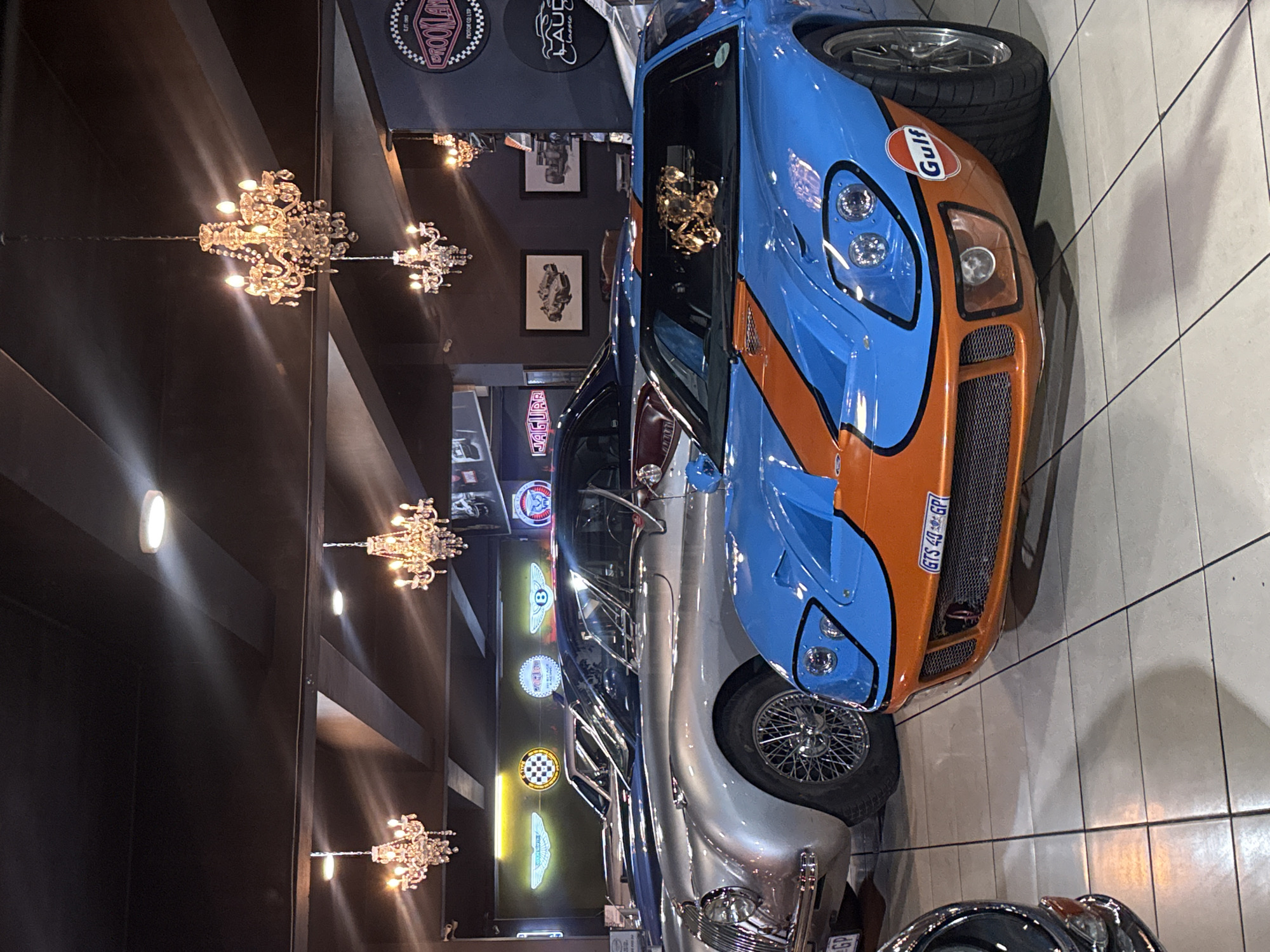 The image depicts the interior of a car showroom located in Cape Town, South Africa. The foreground features a blue and orange Gulf-liveried sports car, partially visible, with a South African license plate ("GTS 40 GP"). Behind it, partially obscured, is a silver vintage car, and in the background, several other classic cars are lined up. The showroom is decorated with vintage car memorabilia, including framed pictures on the wall and illuminated car logos (Jaguar, Bentley). Chandeliers hang from the dark ceiling, adding a touch of luxury. The floor is tiled. There are no people visible in the shot.